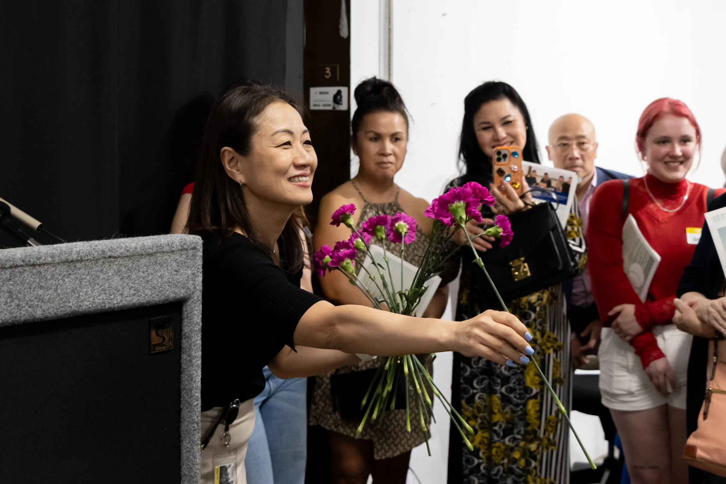 A woman is handing a bouquet of purple flowers to someone else in a group setting, while several people watch and take photos.