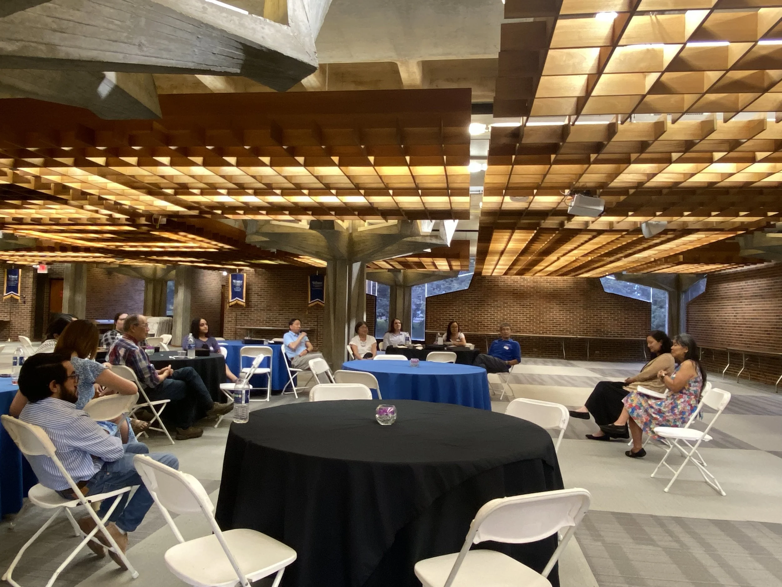 People seated at round tables in a conference room, listening to a speaker. The room has a modern wooden grid ceiling, brick walls, and large windows.