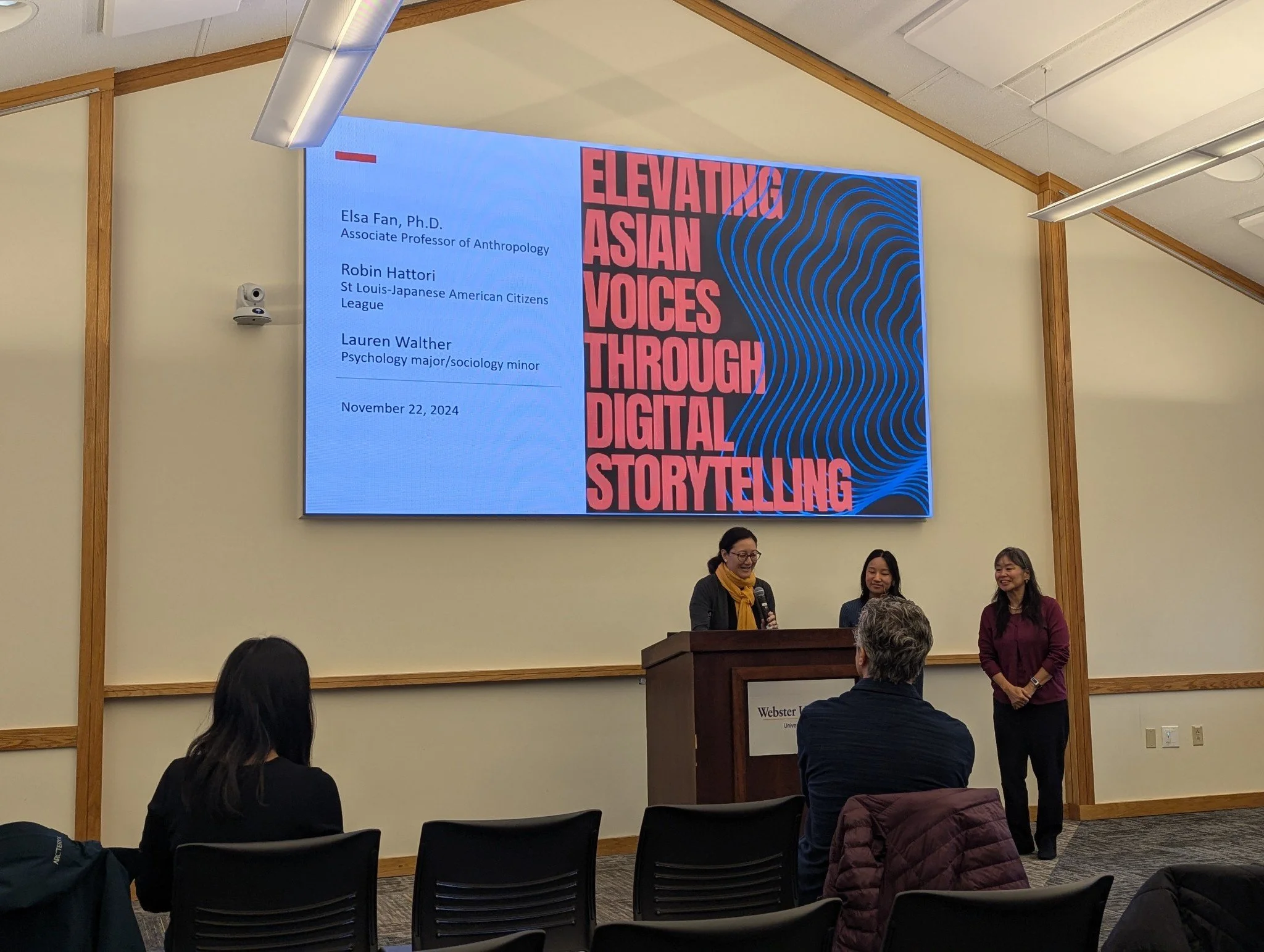 Three women speaking at a podium in front of an audience in a classroom or conference room with a large digital screen behind them. The screen displays a presentation titled 'Elevating Asian Voices Through Digital Storytelling' with names and date listed.