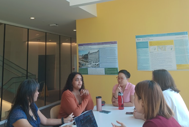 Five young women sitting around a white table in a bright room, engaging in a discussion. Two have laptops open, two have water bottles, with posters on a yellow wall behind them.