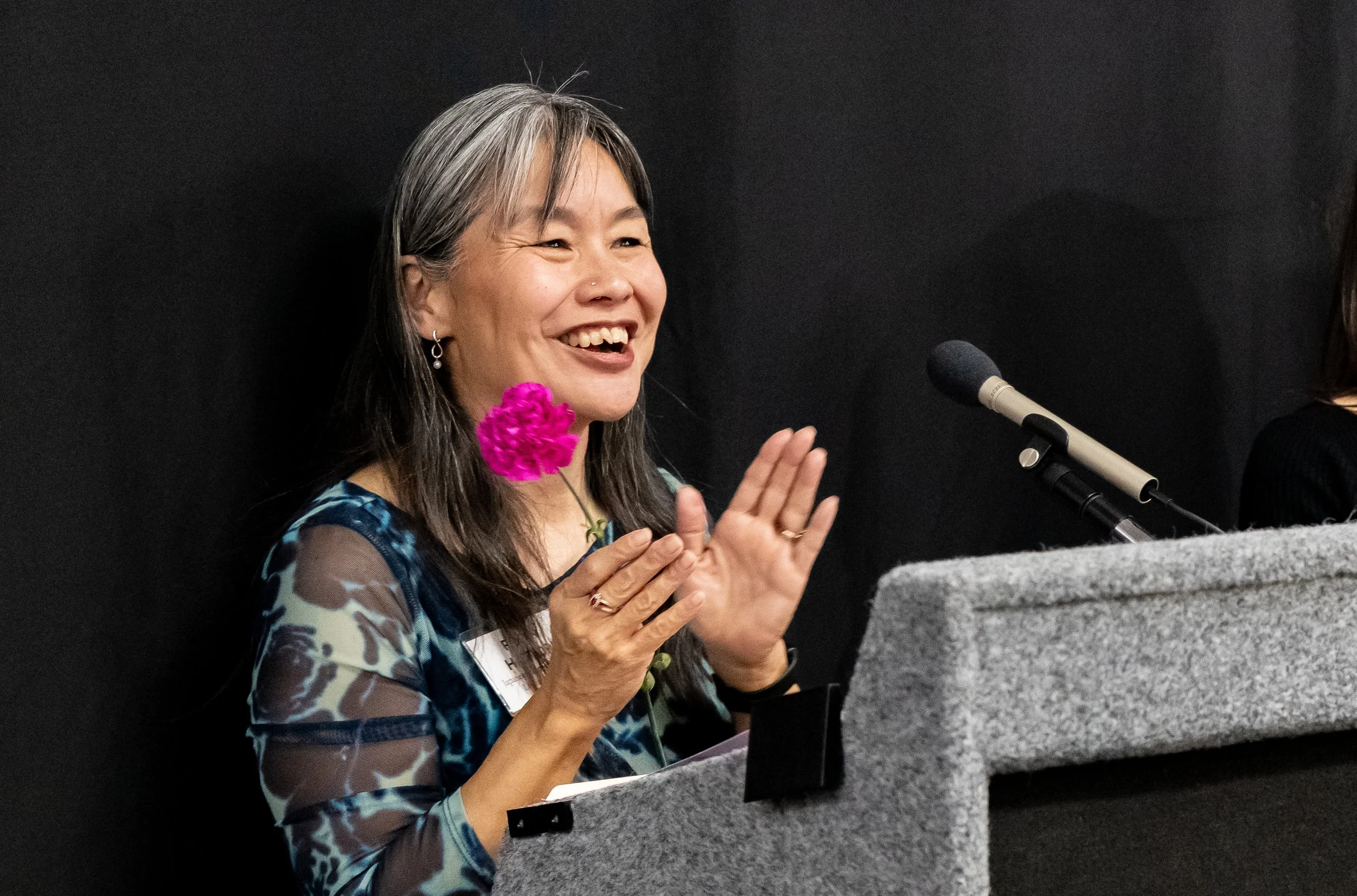 A woman with gray hair, wearing a blue patterned top and earrings, is smiling and clapping. She holds a pink flower in one hand and is seated at a grey podium with a microphone in front of her.