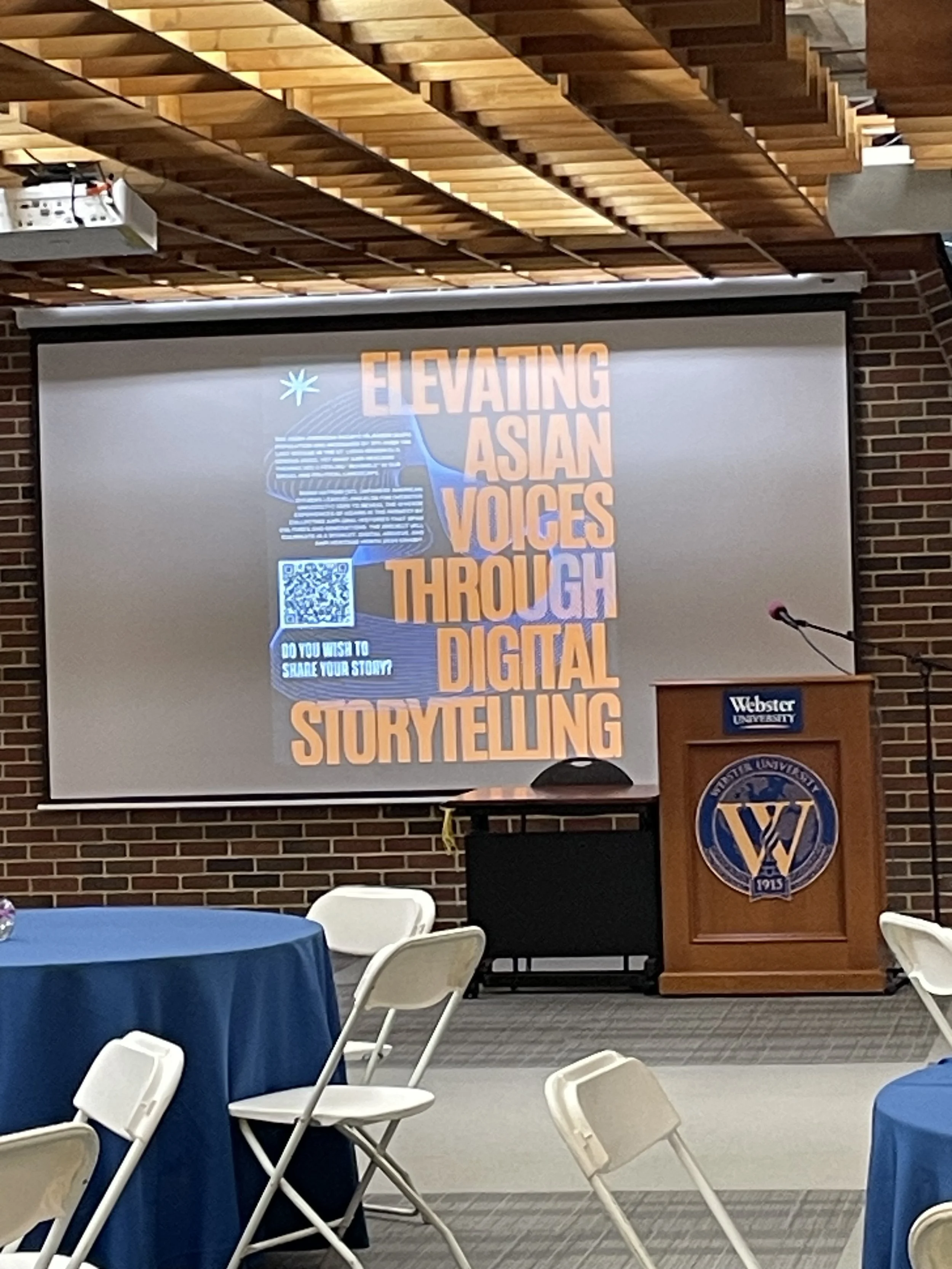 A presentation slide with the text 'Elevating Asian Voices Through Digital Storytelling' displayed on a projector screen in a room with a brick wall. There are tables with blue tablecloths and white chairs in the foreground, and a podium with the Webster University logo on it.