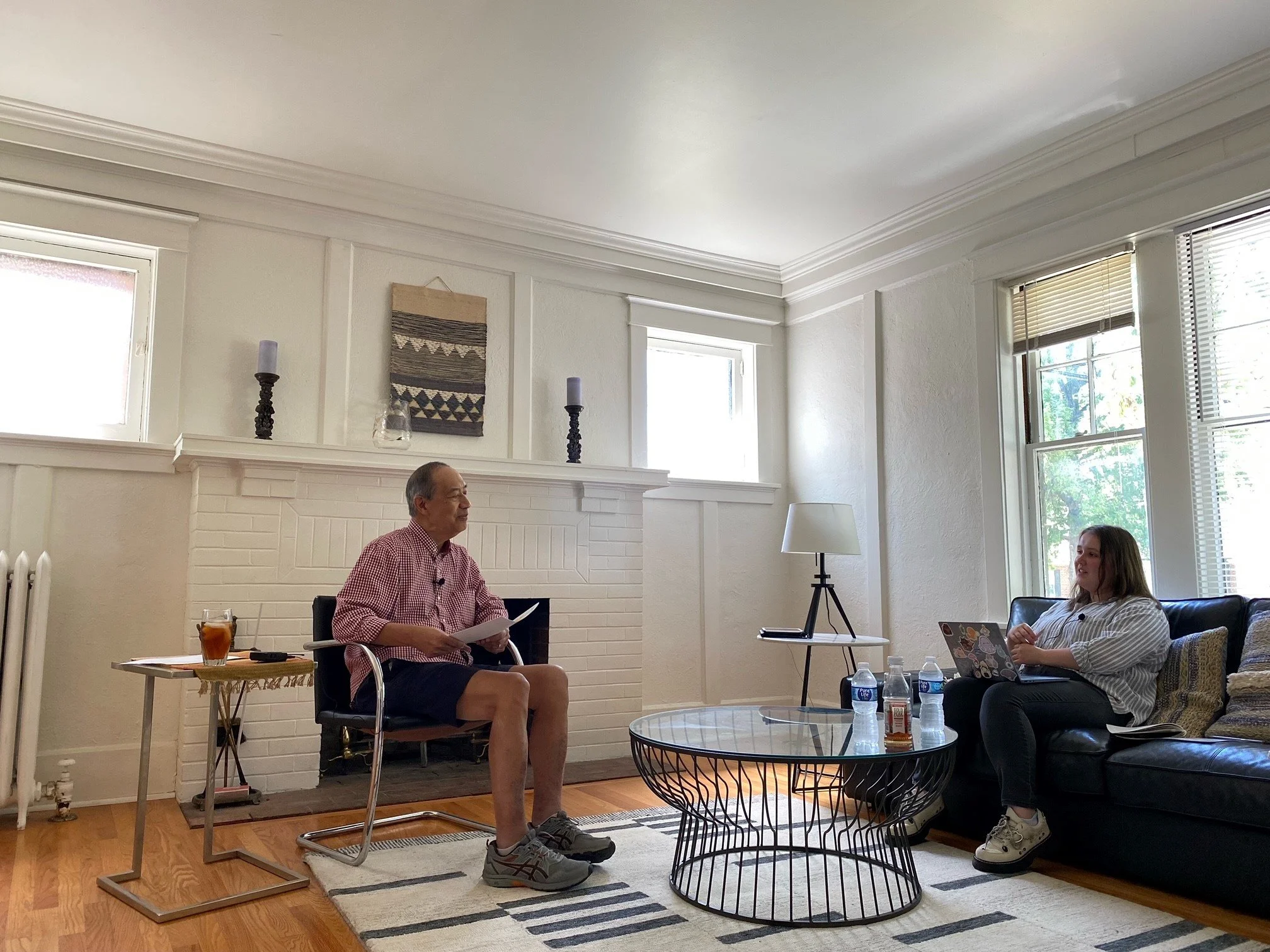 A man and a woman in a living room having a conversation. The man is sitting on a chair near the fireplace, holding papers, wearing a red checkered shirt and shorts. The woman is sitting on a black sofa with a laptop on her lap. There is a coffee table with water bottles and snacks between them and a side table with a drink near the man. The living room has large windows with blinds, a white brick fireplace, and decorative items on the mantle.
