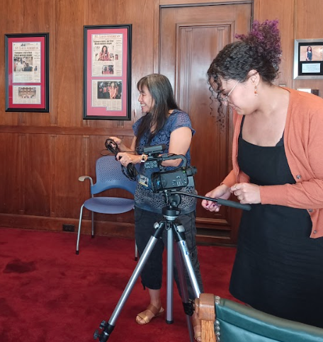 Two women documenting or photographing an event in a wood-paneled room with framed posters on the wall. One woman operates a camera on a tripod, and the other holds a device, possibly a tablet or phone.