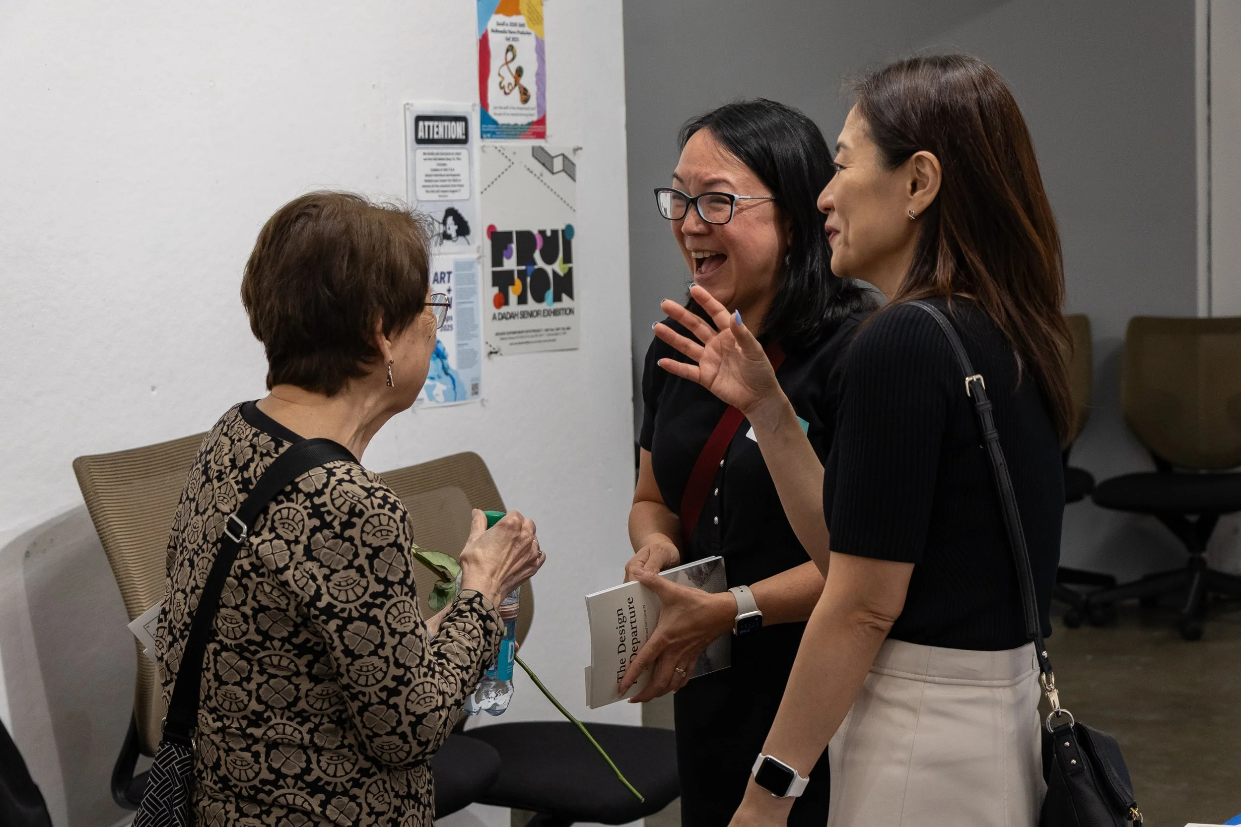 Three women engaged in conversation indoors, with posters on the wall behind them, one of which has the word 'TRU' in colorful letters and mentions an art exhibition.