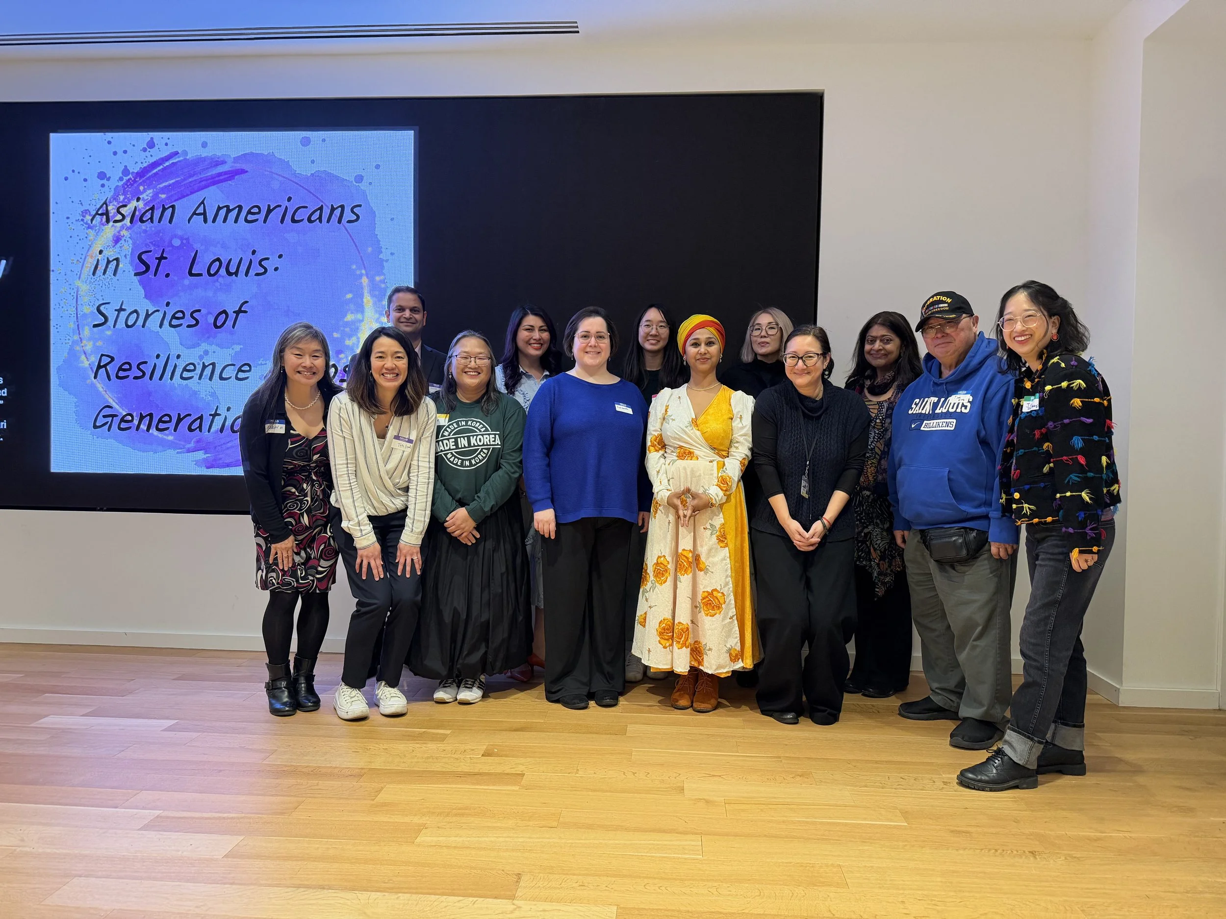 Group of diverse Asian Americans at a presentation or event, standing in front of a screen displaying the title 'Asian Americans in St. Louis: Stories of Resilience and Generations.'
