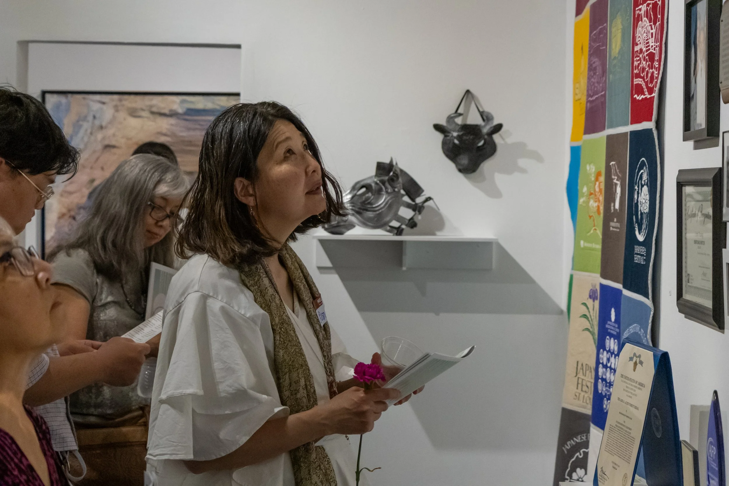Group of people viewing artwork at an art gallery, with a woman in white holding a flower and reading a booklet.