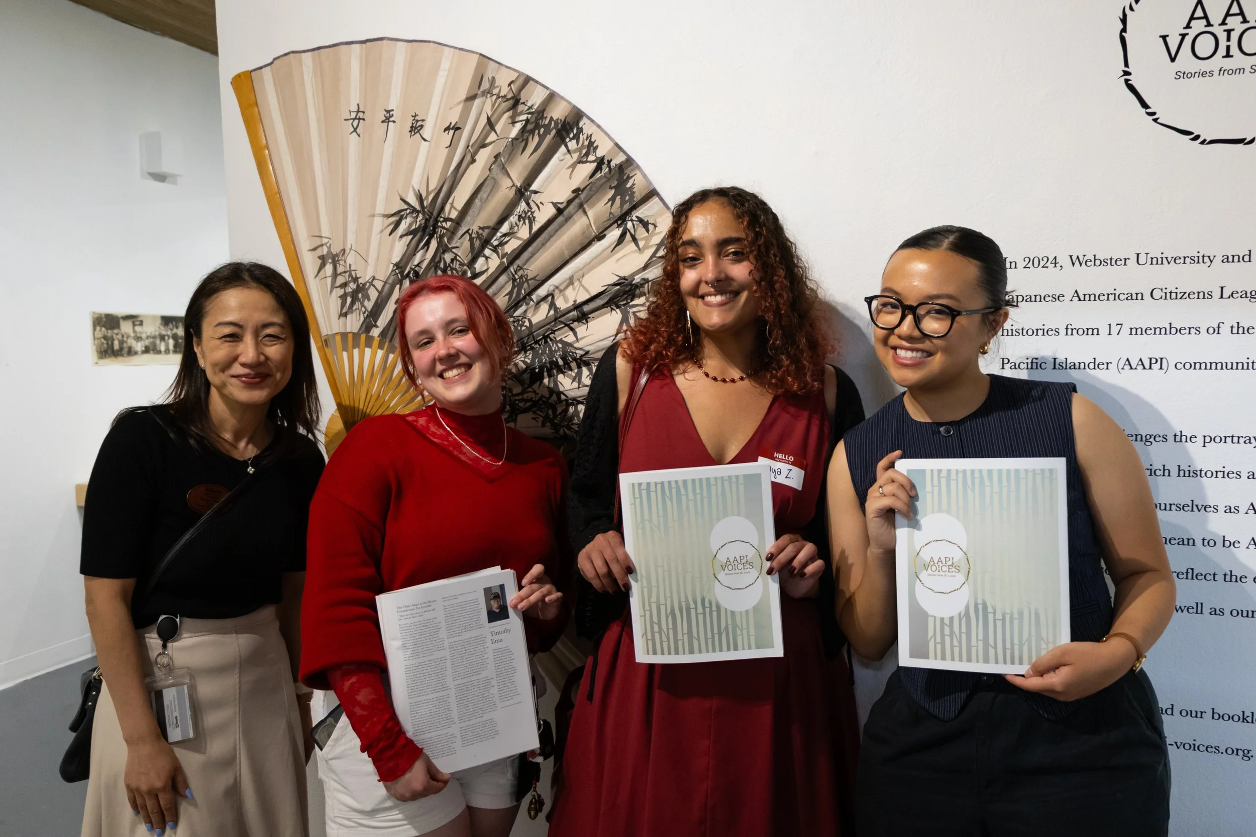 Four women standing together at an event, with two of them holding certificates. They are smiling and posing in front of a wall with a large decorative fan with bamboo and Japanese characters, and a partial wall display.