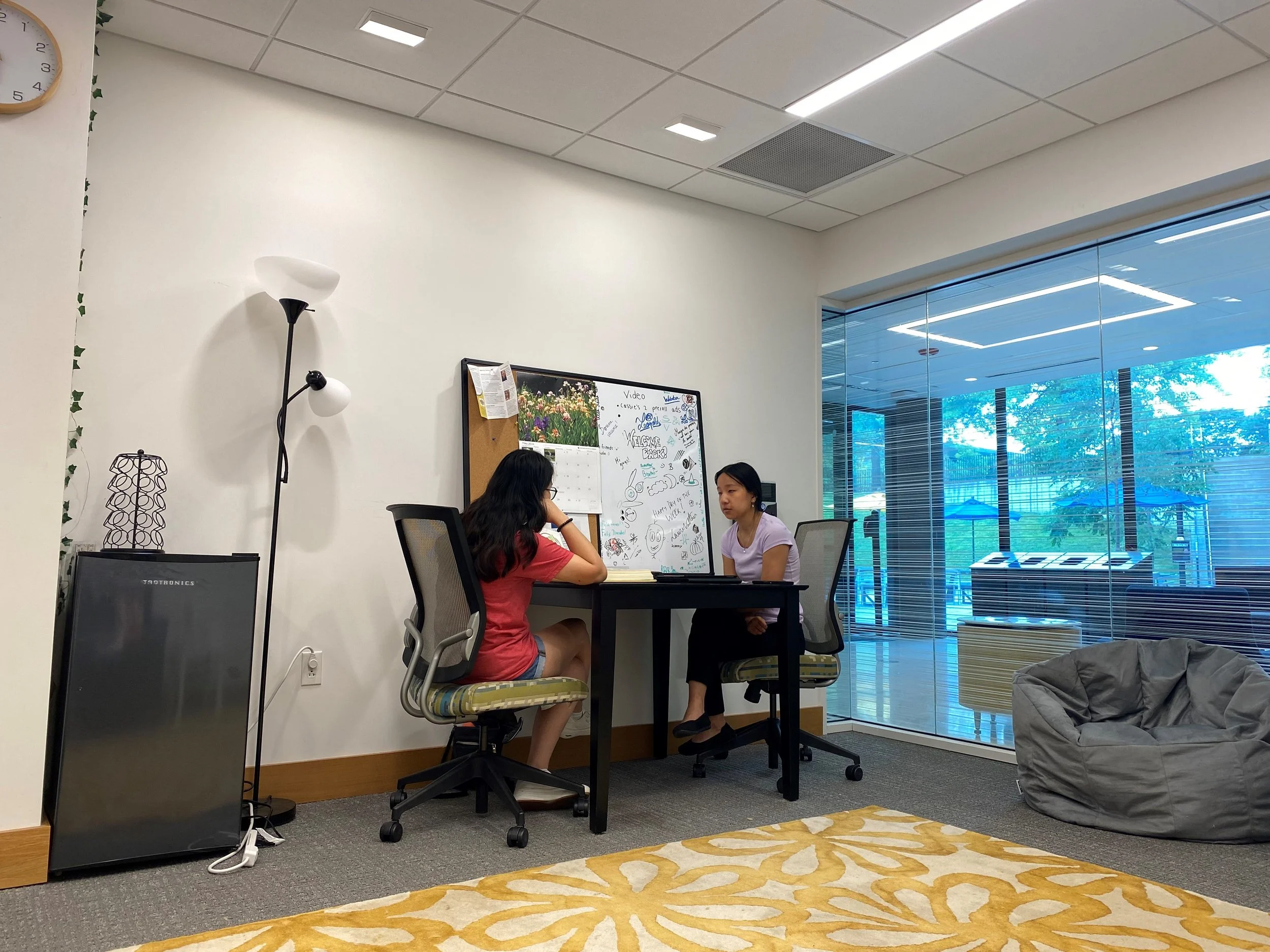 Two women sitting across from each other at a table in an office or lounge area, engaged in conversation. One woman is wearing a red shirt and denim shorts, the other is wearing a white shirt and black pants. The background shows a whiteboard, a large window with a view of trees and blue umbrellas outside, and a bean bag chair. There is a floor lamp, a mini refrigerator, and a decorative lamp nearby.