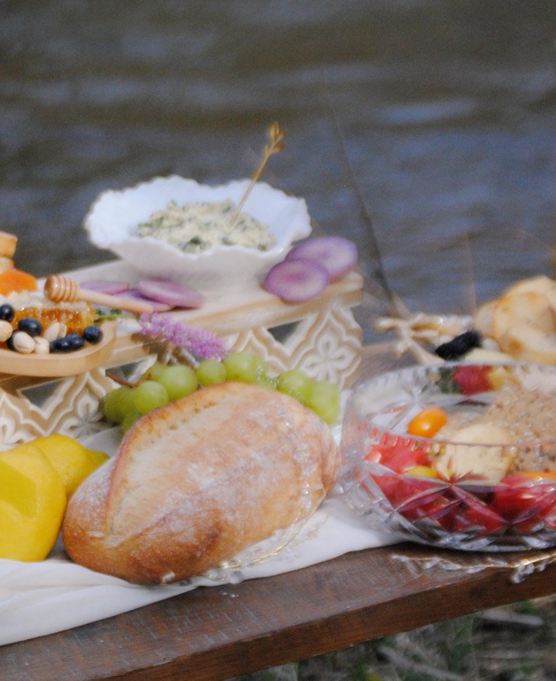 A rustic spread of food including a loaf of bread, grapes, cheese, fruit salad, and a bowl of dip on a wooden table near water.