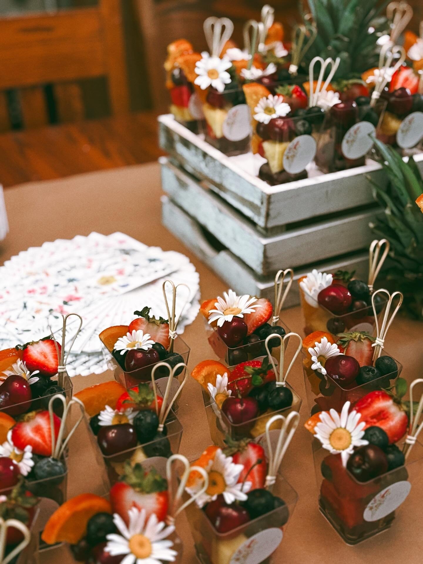 Dessert cups filled with strawberries, blueberries, cherries, and orange slices, decorated with small daisies and white decorative sticks, arranged on a table with a wooden and white crate backdrop.