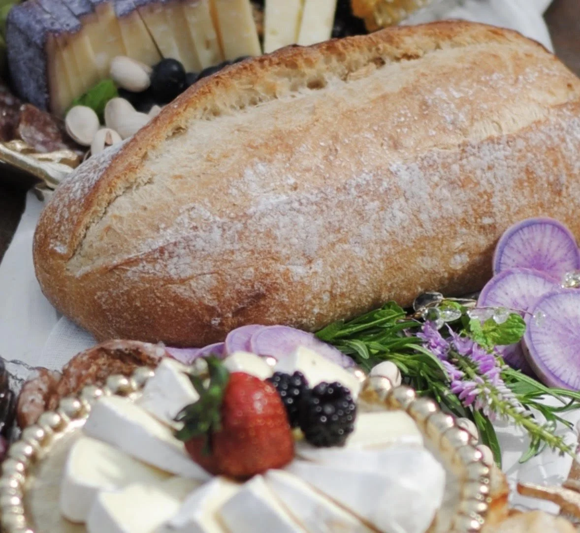 A dessert display with a glass dome covering a layered cake decorated with strawberries and blackberries, surrounded by assorted cookies, nuts, strawberries, and small jars of jam or syrup.