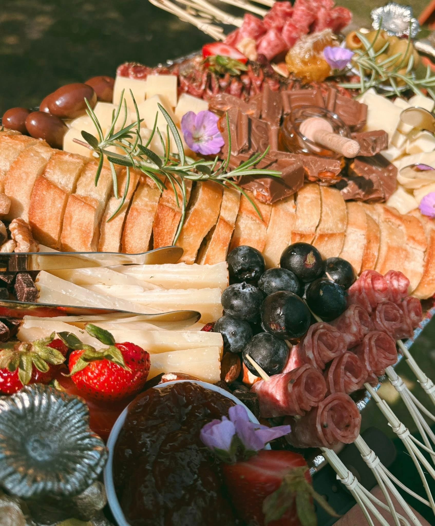 A cheese, fruit, and chocolate platter with strawberries, blueberries, grapes, assorted cheeses, chocolates, and garnished with purple flowers and herbs.