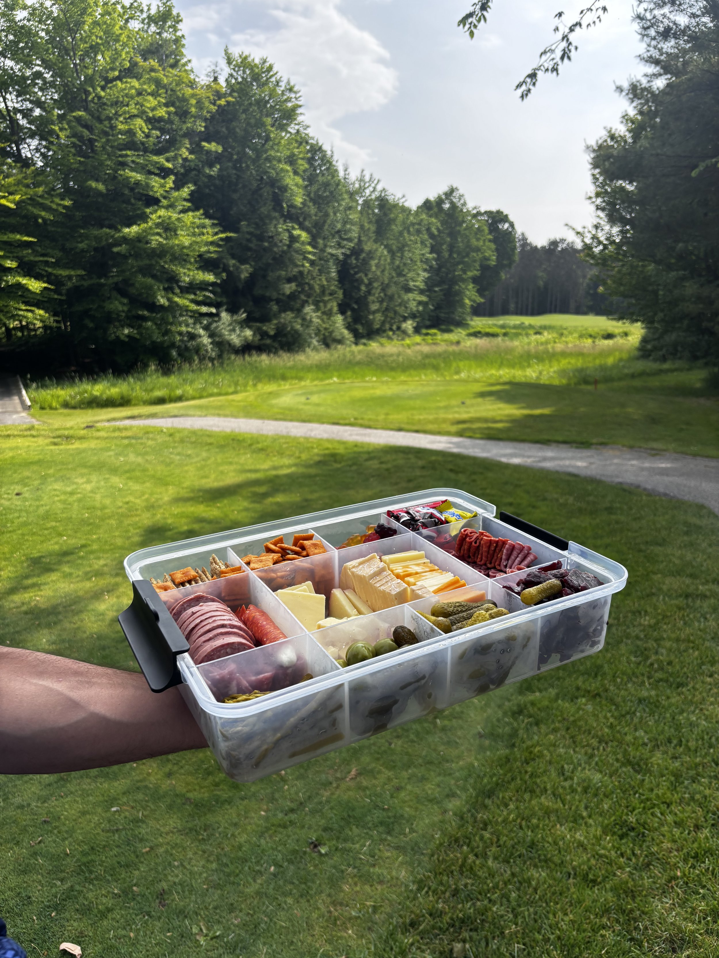 A person holding a plastic food container filled with various cheeses, meats, crackers, and snacks outdoors on a grassy area with trees and a winding path in the background.