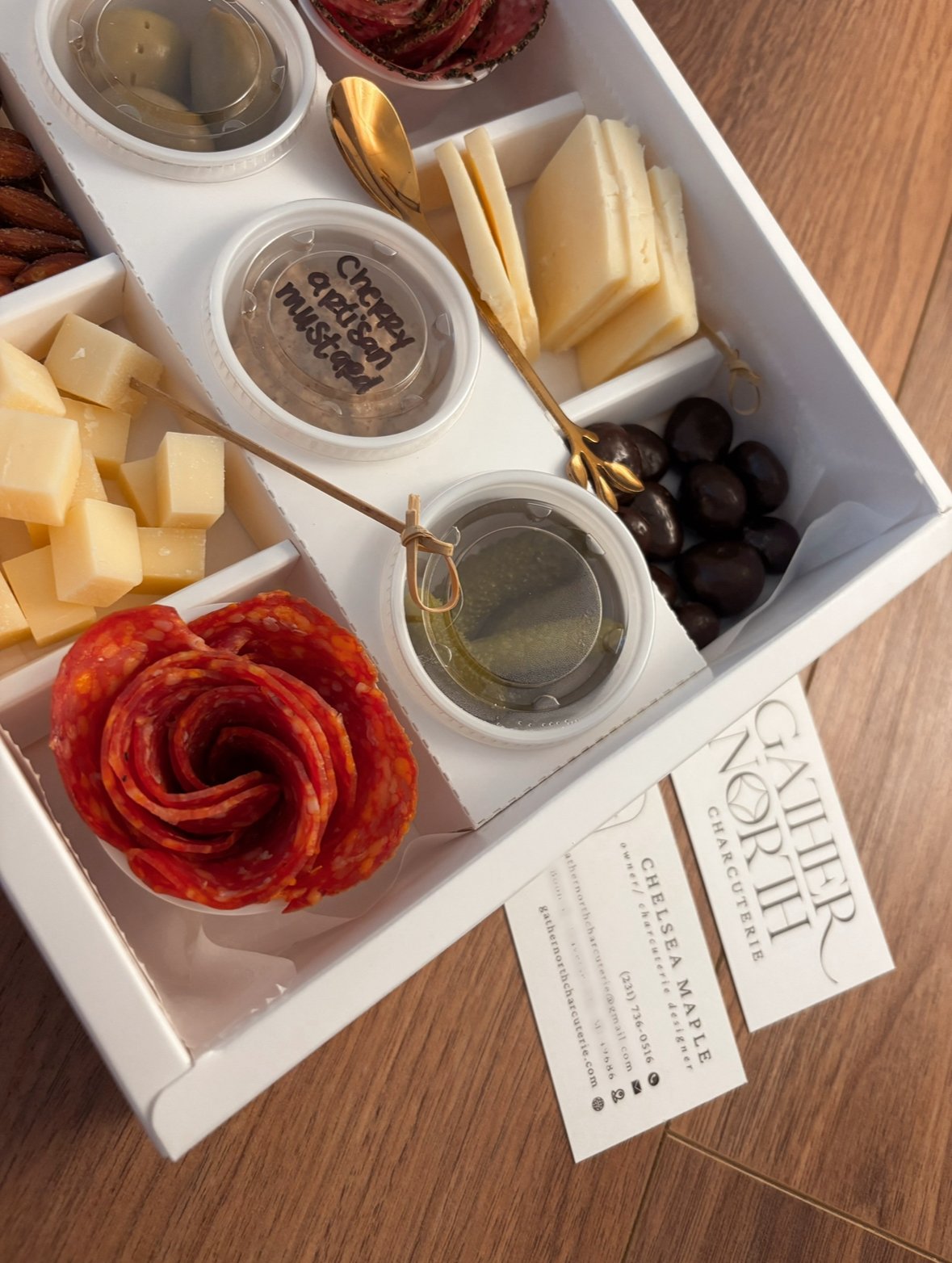A cheese and charcuterie sample tray with various cheeses, slices of cured meat, black olives, green olives, and condiments on a wooden table. There are small containers with labels, one says 'Cheddar', another 'Cherry & Fig Jam', and a third with oi