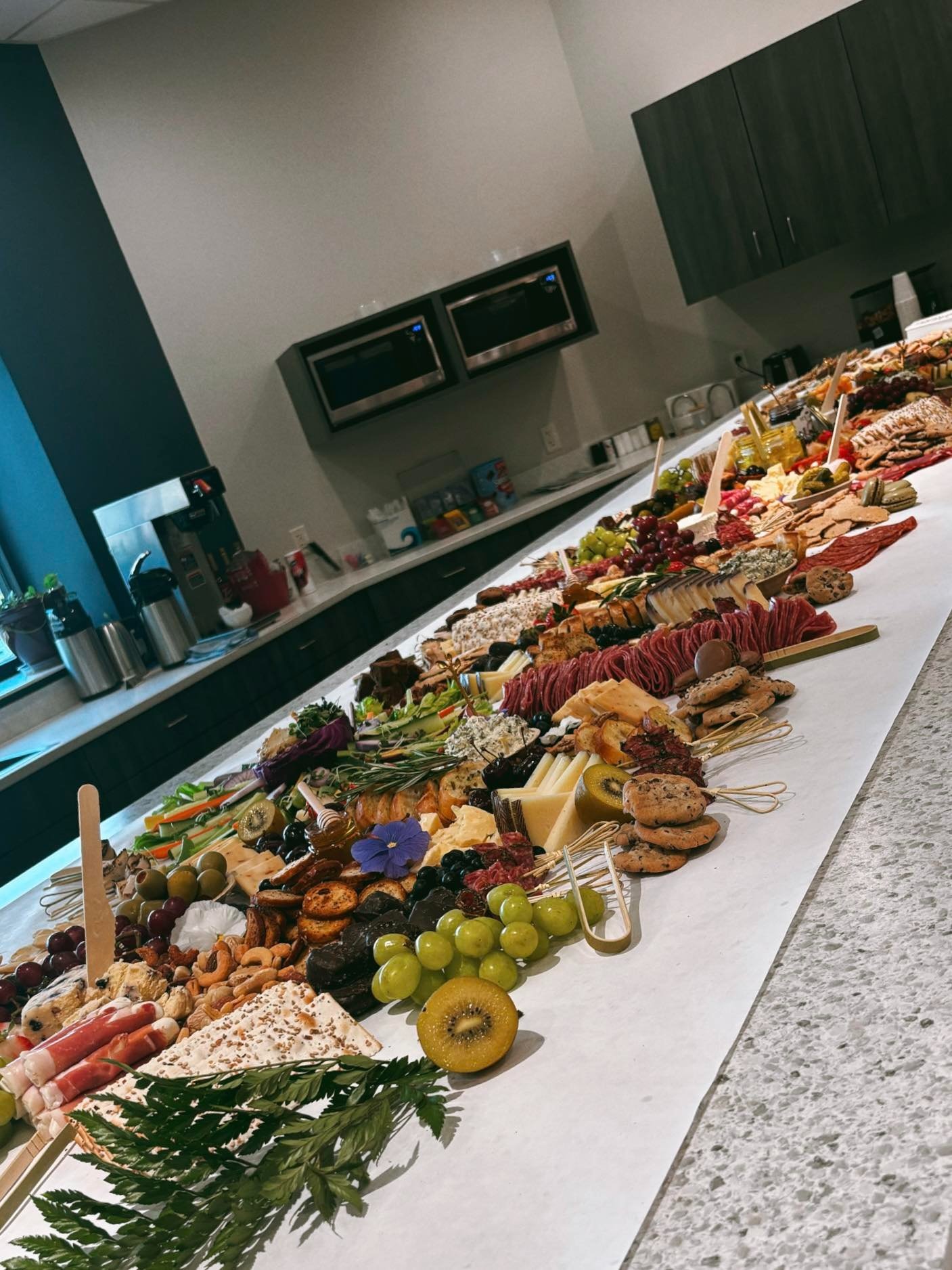 A long table filled with various cheeses, grapes, sliced meats, crackers, and fruit at a buffet. In the background, there are kitchen cabinets, microwaves, and coffee pots.