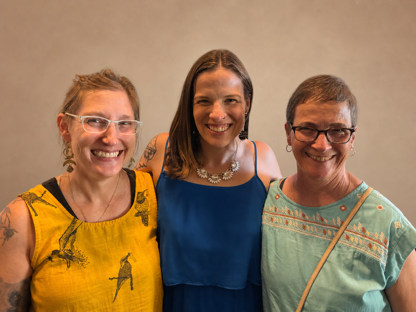 Three women smiling in front of a plain wall. The woman on the left has short blonde hair, glasses, and tattoos, and is wearing a yellow top with black patterns. The woman in the middle has long brown hair, earrings, and a necklace, and is dressed in a blue sleeveless top. The woman on the right has short gray hair, glasses, and hoop earrings, and is wearing a light green top with embroidery.