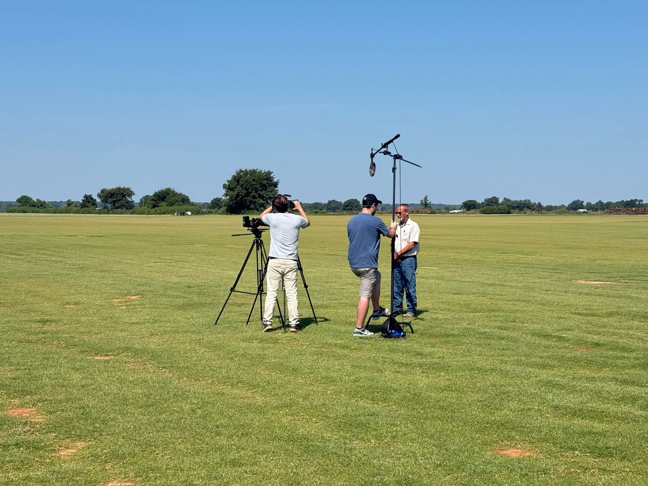 Three people standing in an open field, conducting an interview or recording session with a boom microphone and camera, under a clear blue sky.