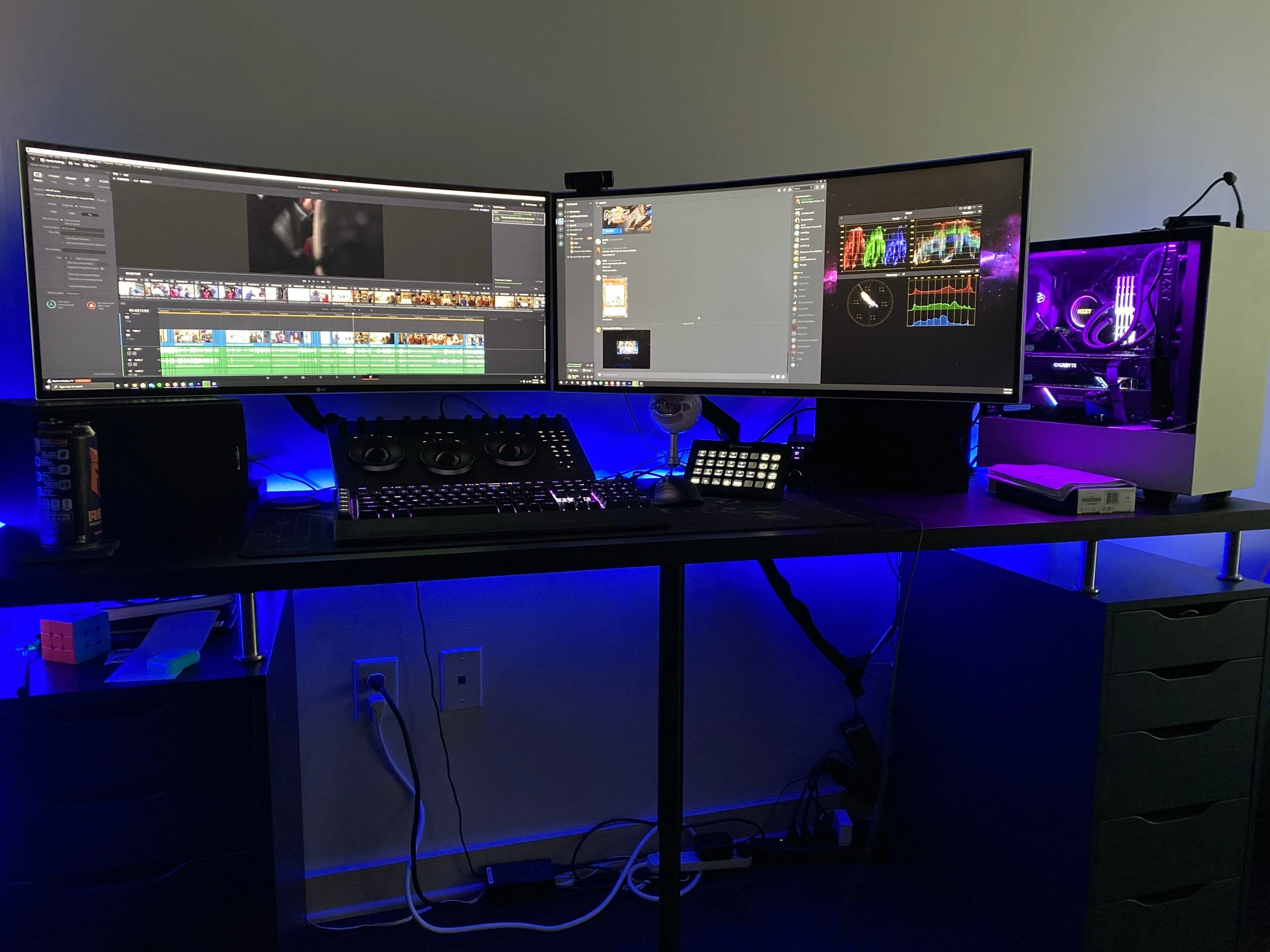 A dual-monitor computer setup on a desk, with a desktop tower featuring purple lighting on the right side. The monitors display video editing and communication applications, with a keyboard, mouse, and various peripherals on the desk. There is a can of soda on the left and some small objects on the desk.