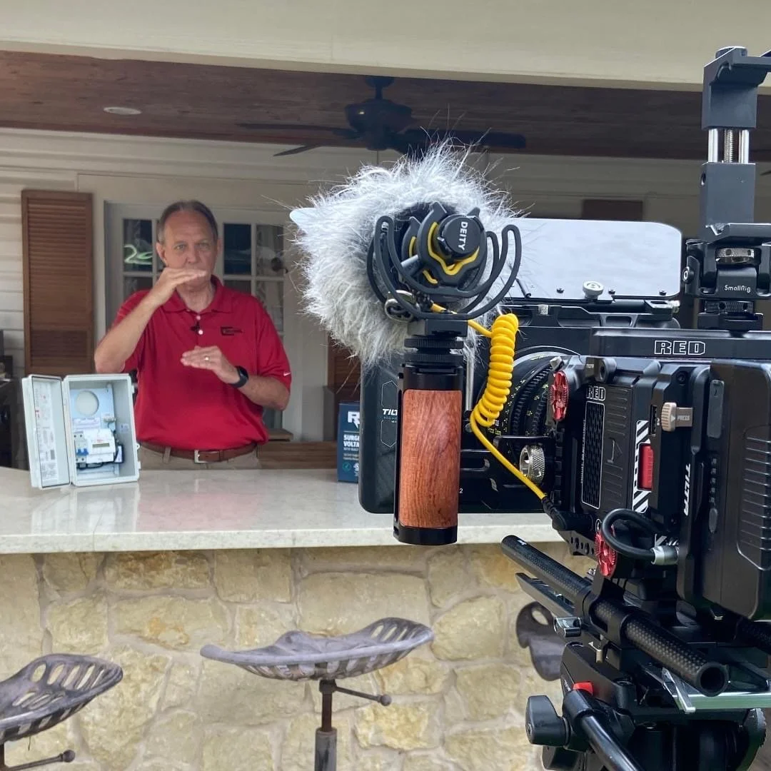 A man in a red polo shirt standing behind a counter, speaking or explaining, with professional filming equipment and a camera in the foreground, outdoors on a porch or patio area.