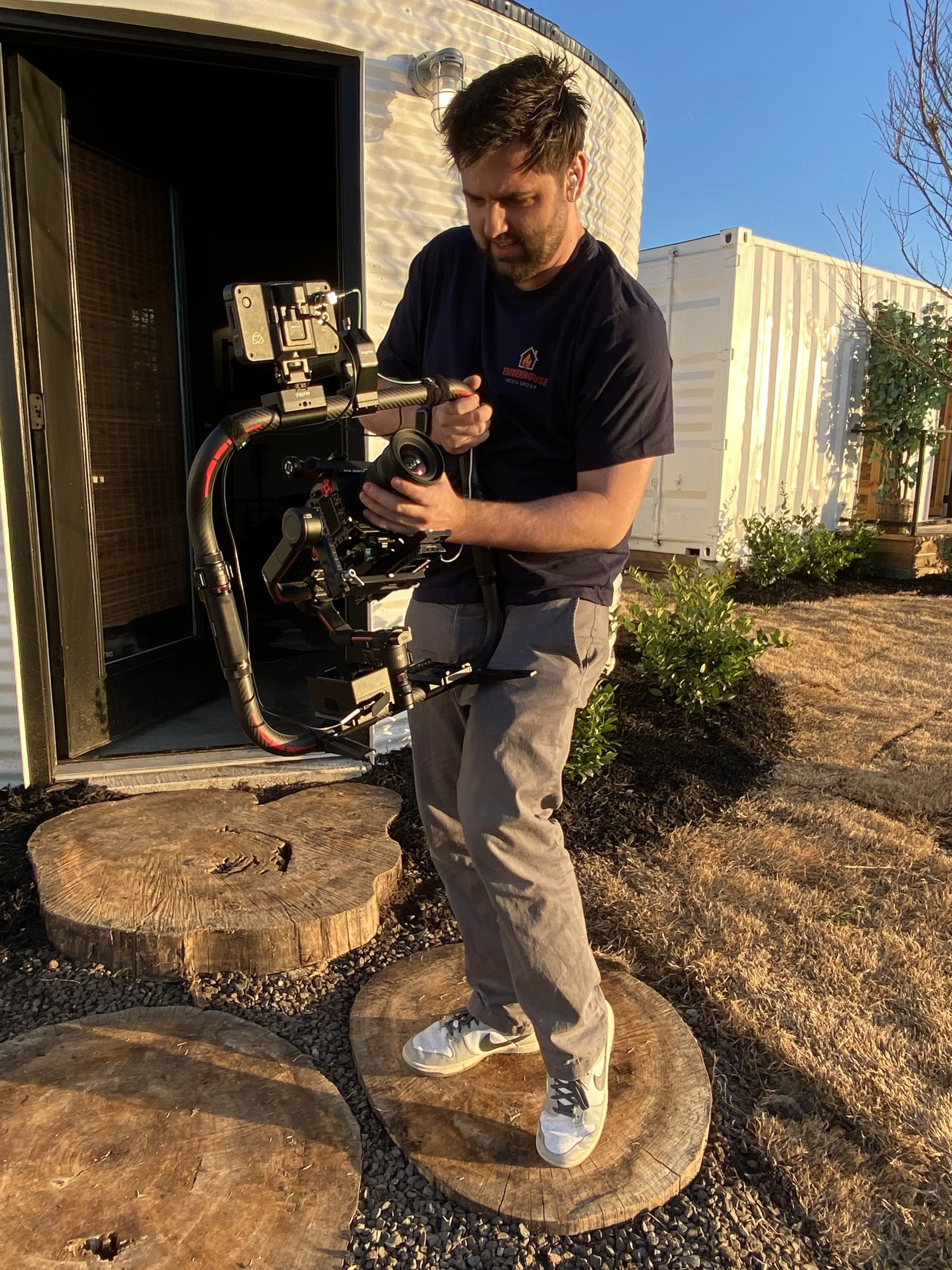 A man wearing a black t-shirt and gray pants stands outdoors on a rocky area, holding a camera stabilizer with a mounted camera. He is looking at the camera with a focused expression. In the background, there are large tree stumps, a white building, and a clear blue sky.