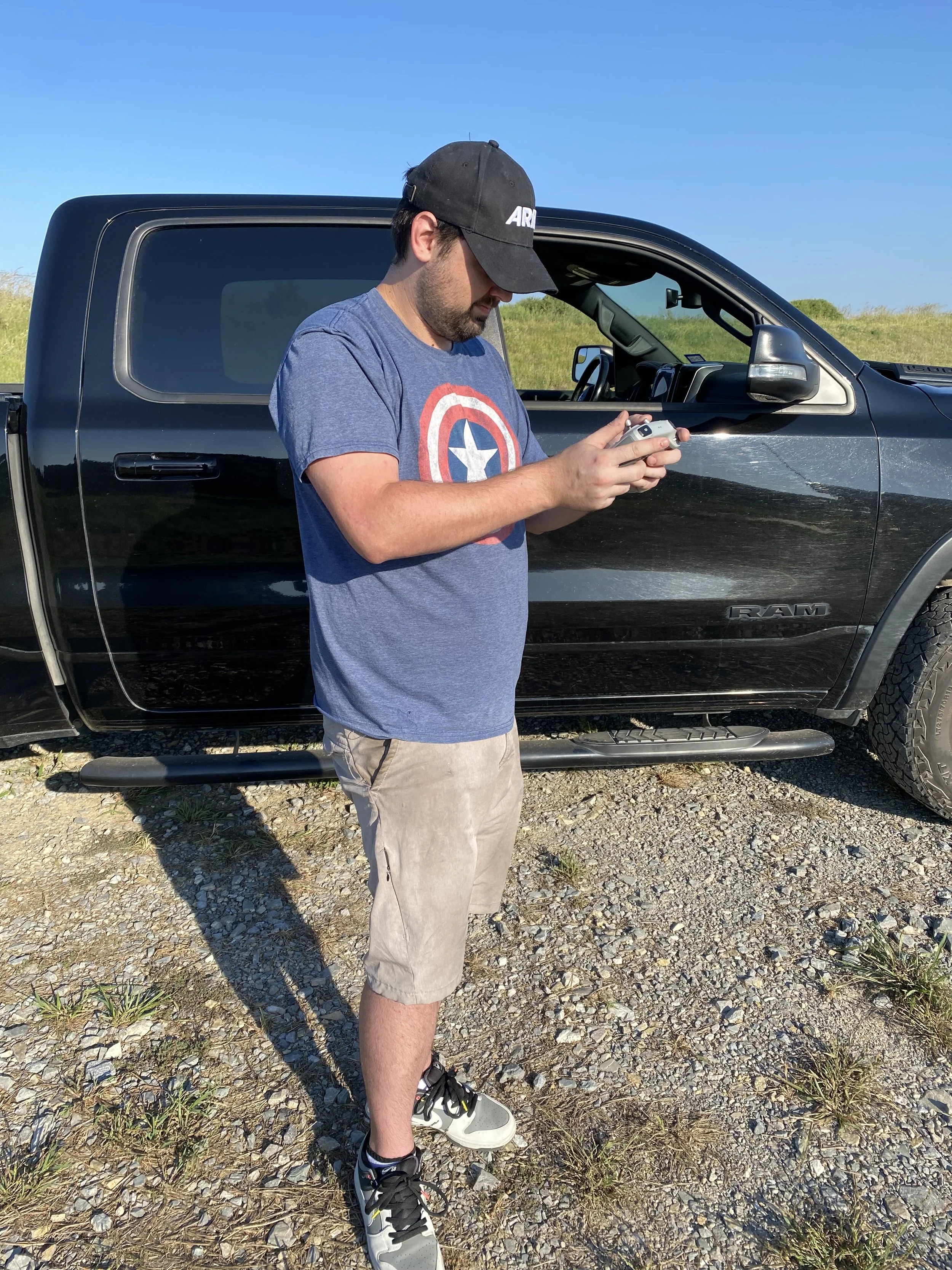 A man wearing a black cap, a blue Captain America t-shirt, beige shorts, and sneakers standing outdoors, looking at his phone with a black pickup truck behind him on a gravel surface with grass and a clear blue sky.