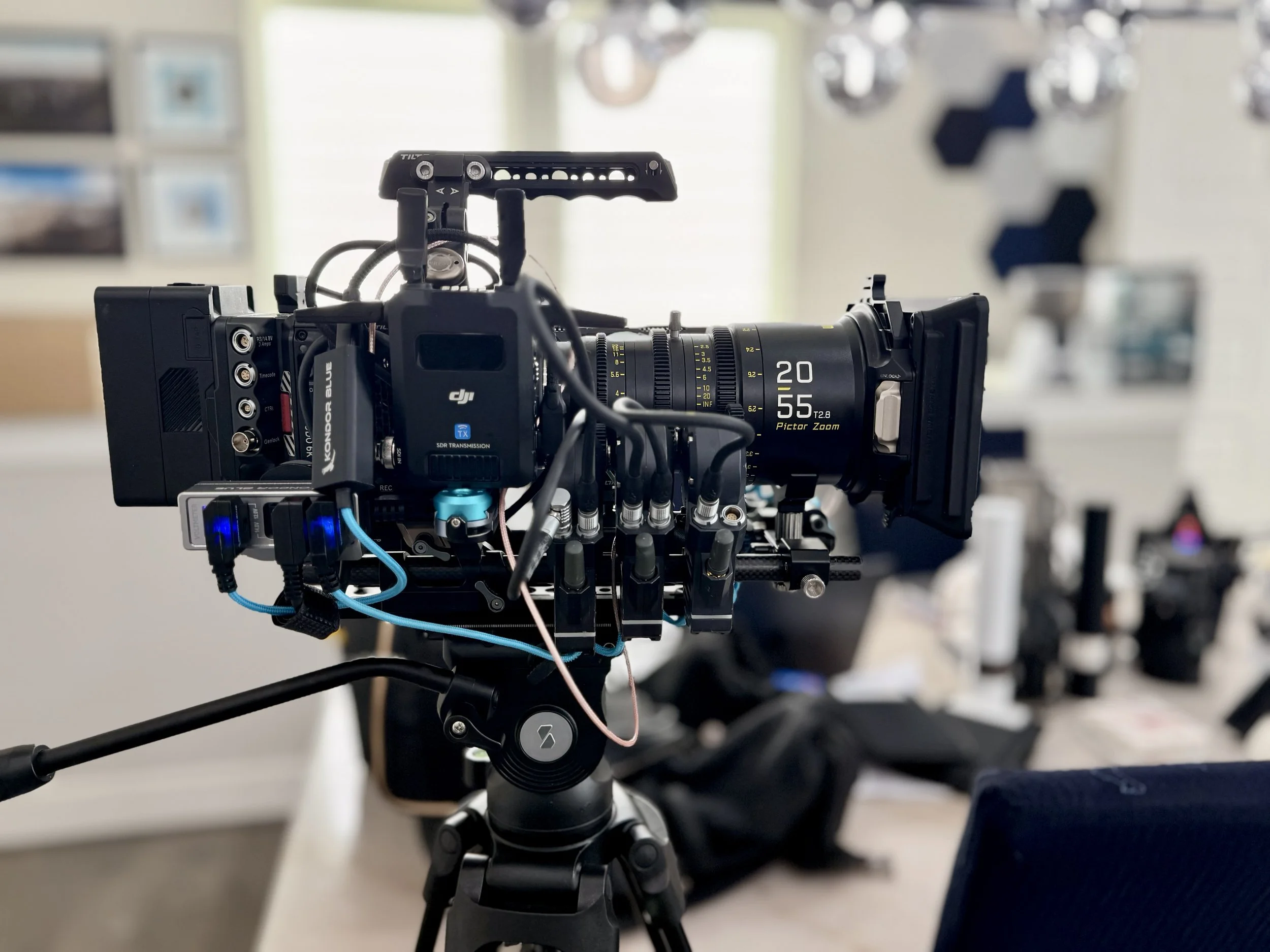 Close-up of a professional camera mounted on a tripod in a room with shelves and blurred objects in the background.