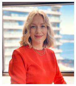 A woman with shoulder-length wavy blonde hair wearing a red top and a necklace, standing in front of a window with a blurred cityscape background.