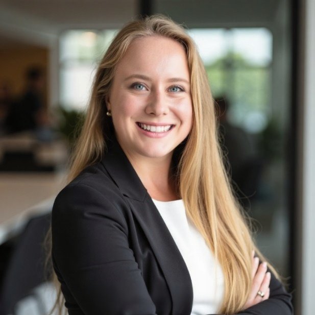 Close-up of a young woman with long blonde hair and blue eyes, smiling, standing outdoors in front of green foliage.