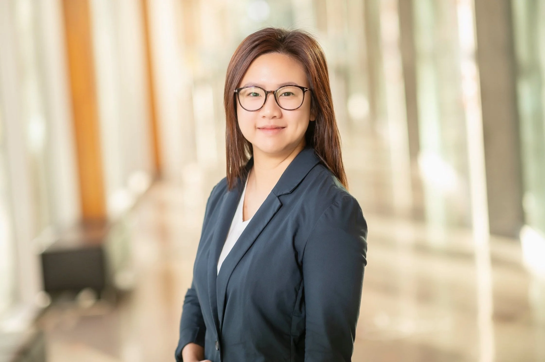 A woman with glasses and shoulder-length brown hair wearing a navy blazer and white top, standing in a well-lit corridor with large windows and trees outside.