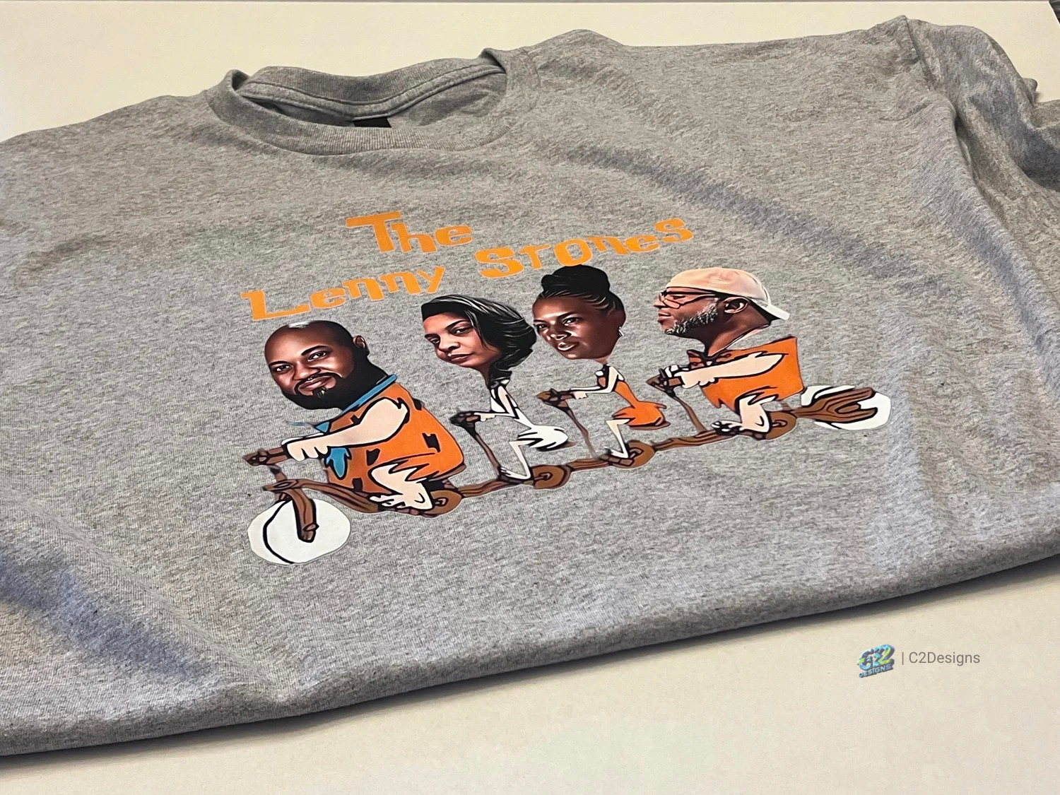 Gray t-shirt with a cartoon spaceship carrying four caricatured human faces and the text "The Lenny Stones" in orange.