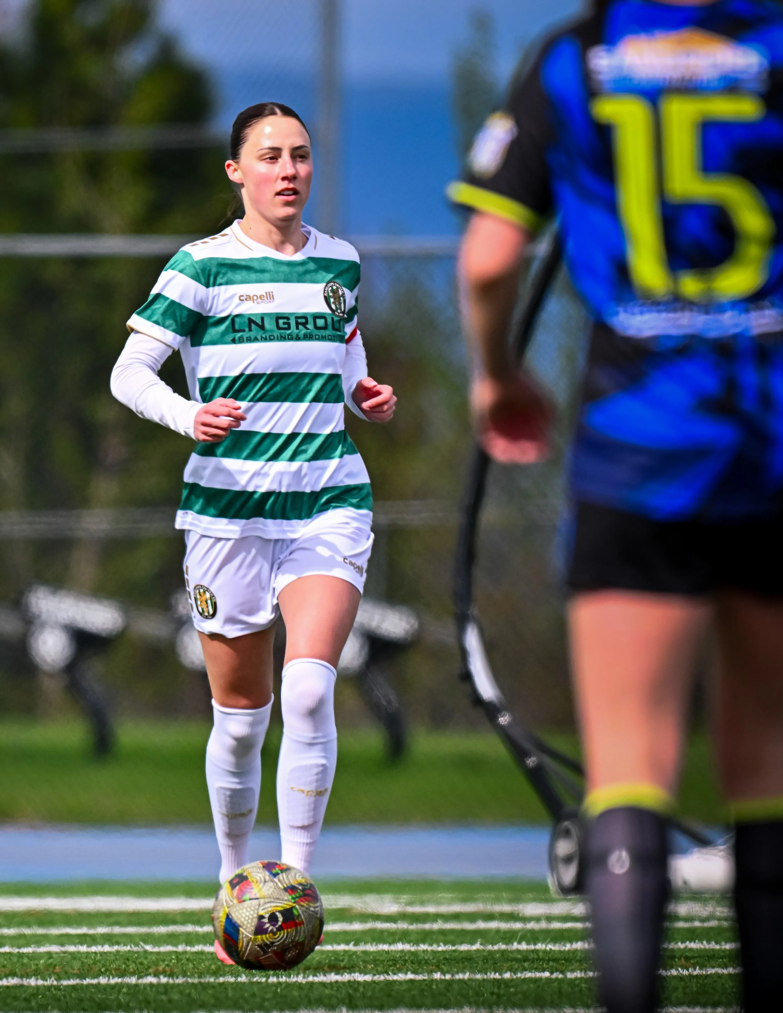 Female soccer player wearing a green and white striped jersey with "CN Group" on the front, dribbling a colorful soccer ball on a field. Another player in a blue and black jersey with a number "15" is partially visible.