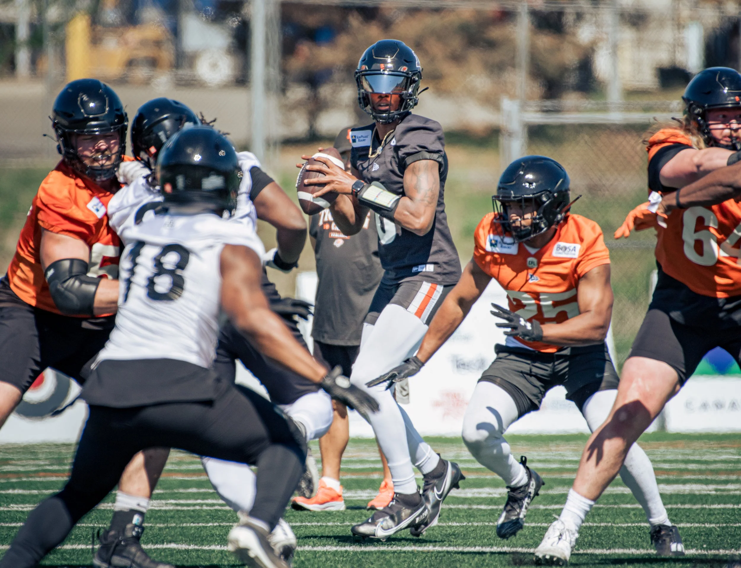 A group of players in football uniforms practicing on a field. One player in black is preparing to throw a football, while others in orange and black jerseys are positioned around him, some blocking, others preparing to receive or tackle. The backgro