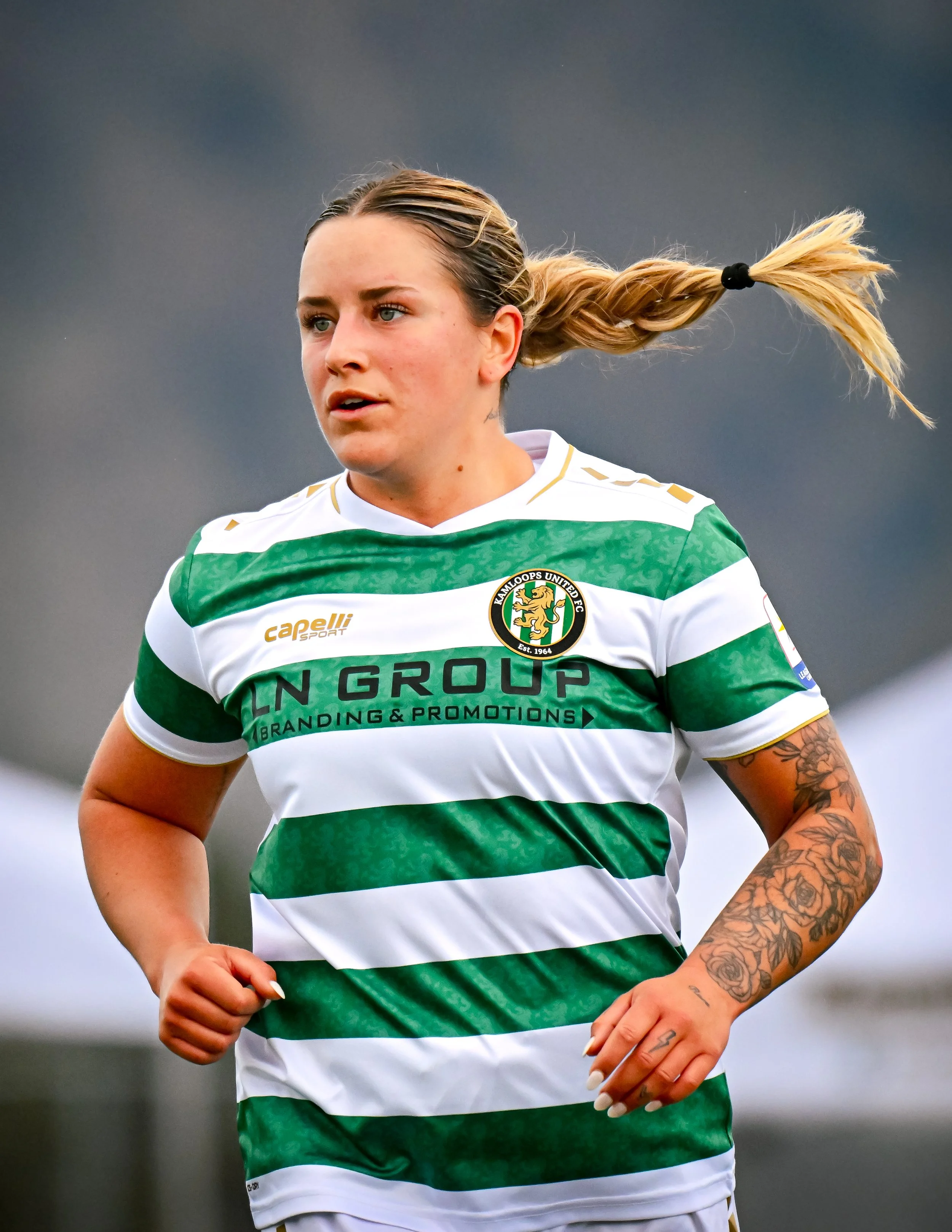 Female soccer player in a green and white striped jersey running during a game, with a tattoo on her arm and her hair in a ponytail.