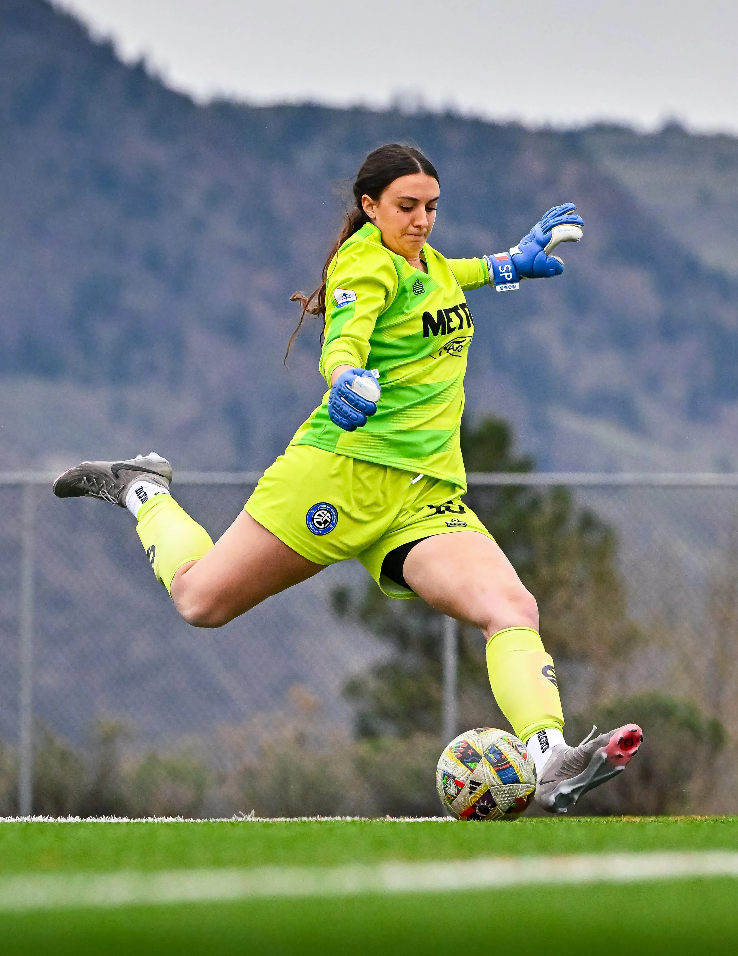 A female soccer player in bright green goalkeeper uniform and blue gloves kicking a soccer ball on a field with mountains in the background.
