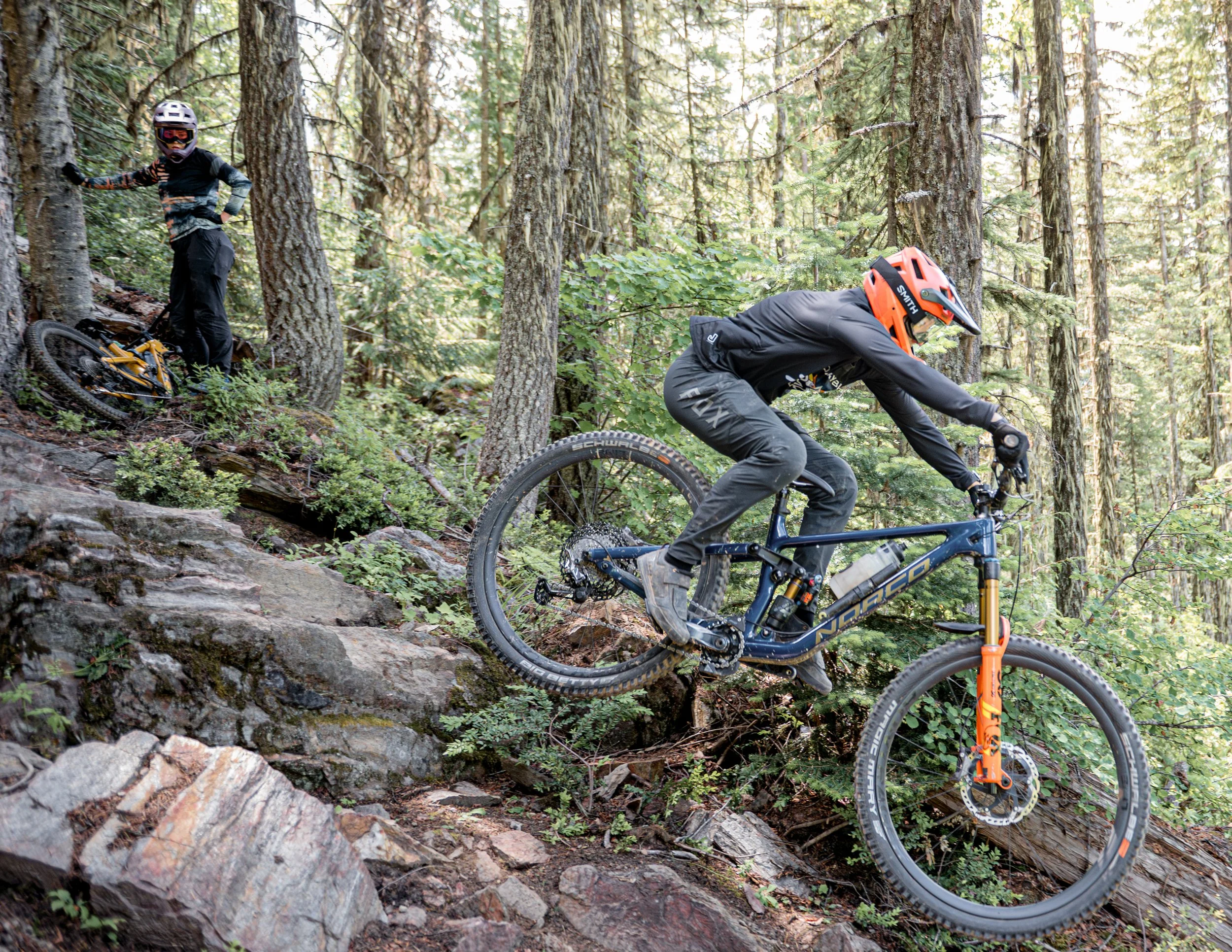 Mountain biker navigating rocky downhill trail in forest with another biker watching nearby.
