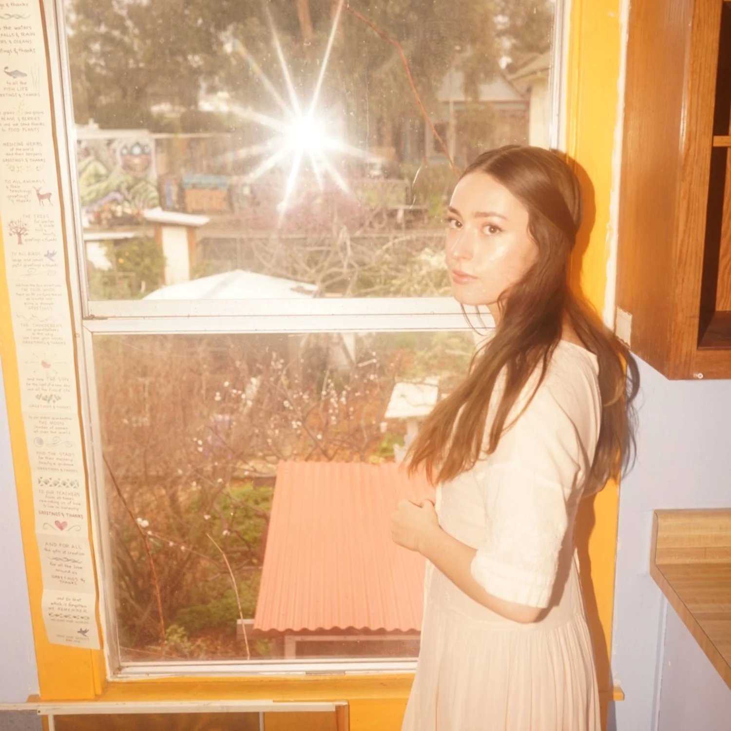 A young woman with long brown hair standing indoors near a window, sunlight shining through, with a garden and trees outside.