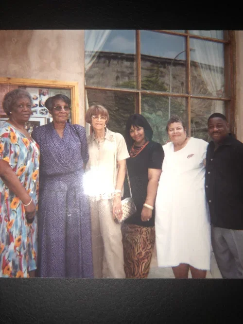 Six women and one man standing indoors in front of a large window, posing for a group photo.