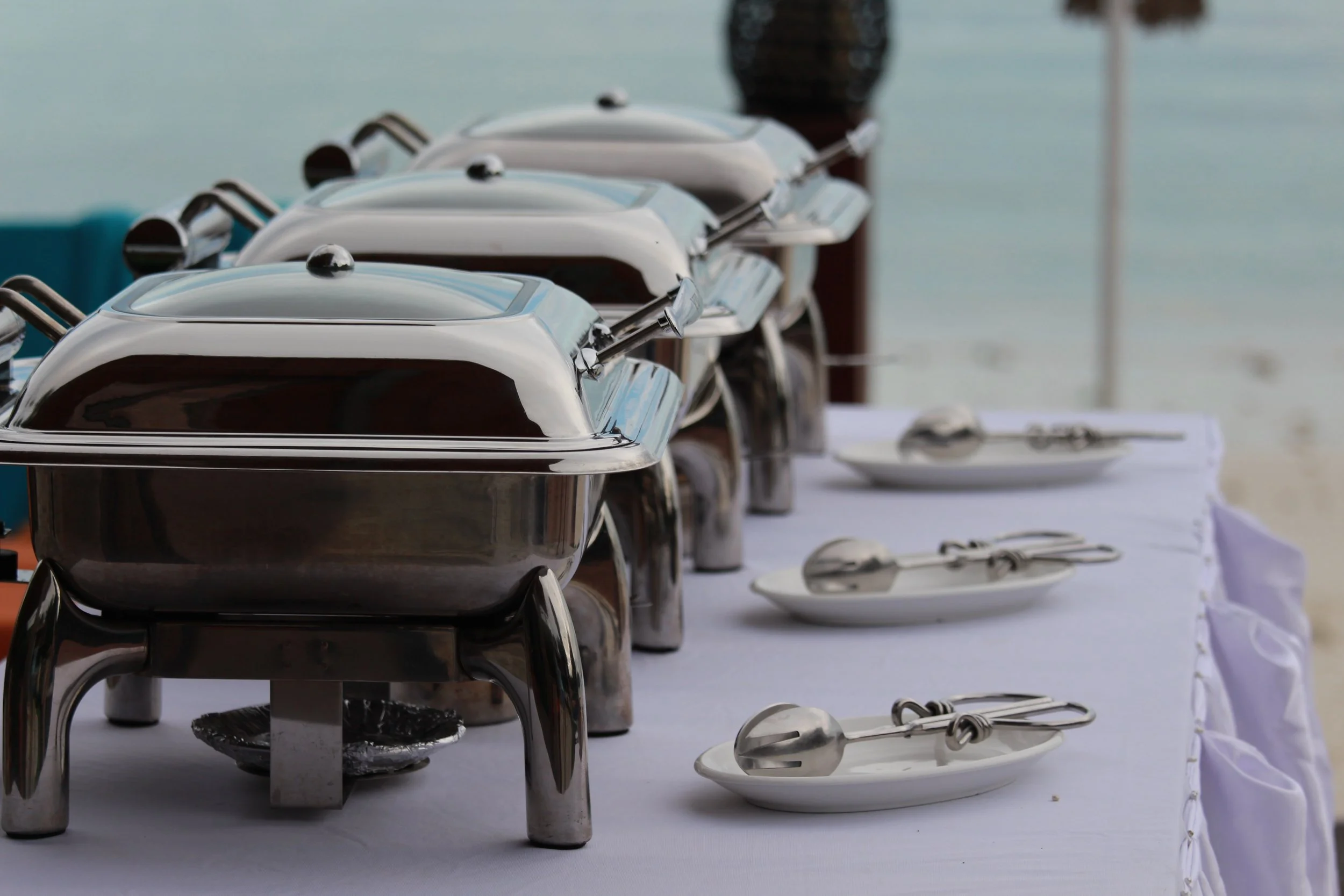 A row of stainless steel chafing dishes and serving spoons on a white tablecloth, with a blurred beach scene in the background.