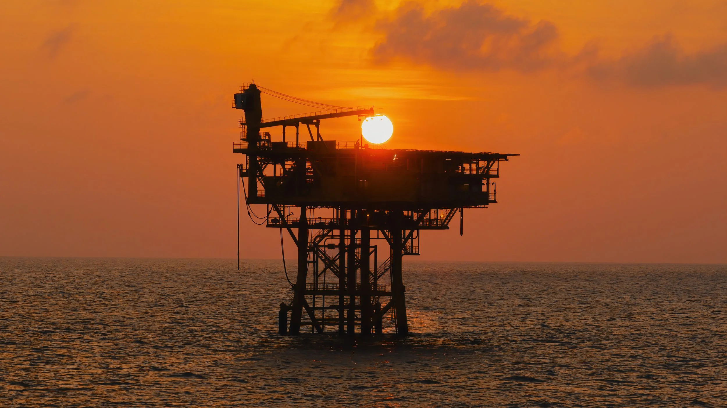 Offshore oil rig silhouette at sunset with the sun setting behind the platform over the ocean.