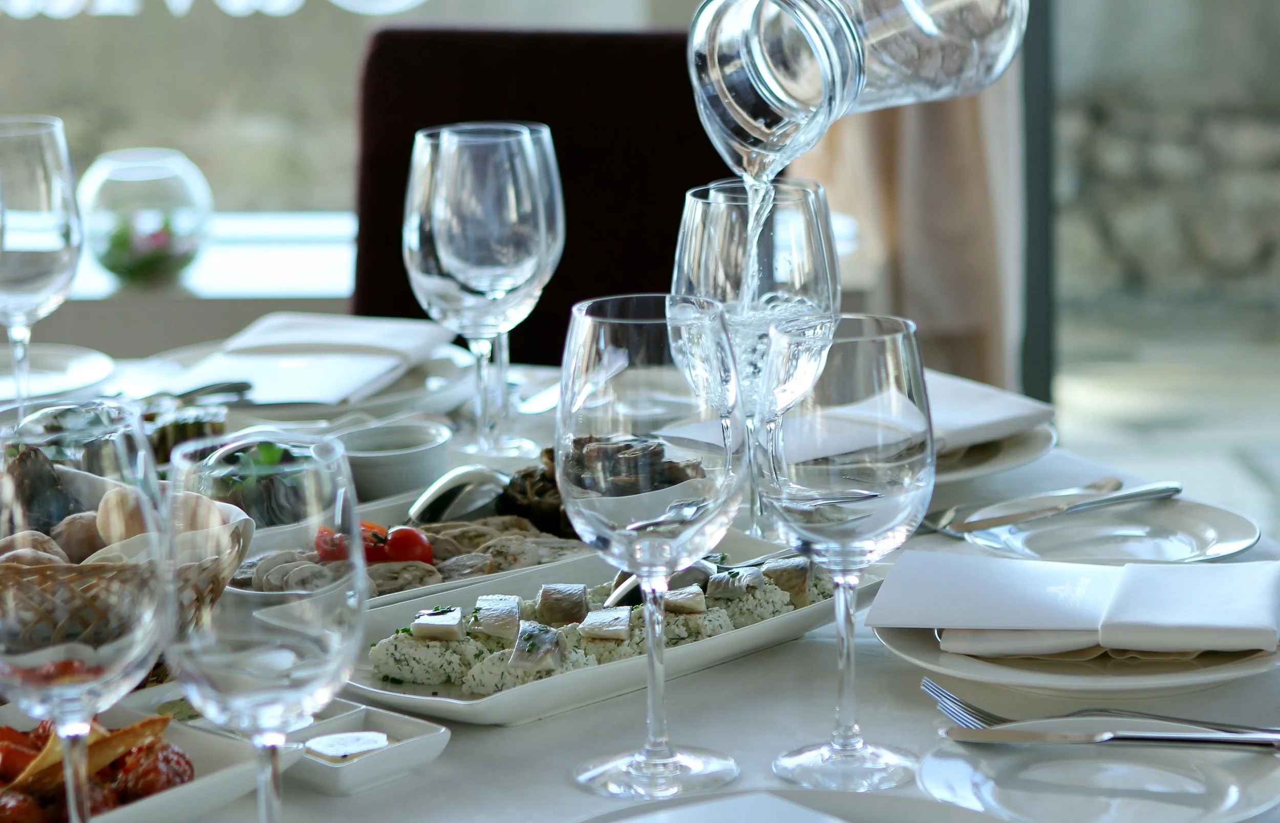 Elegant dining table set with multiple empty wine glasses, plates with white napkins, and a platter of assorted food including cheeses, vegetables, and seafood.