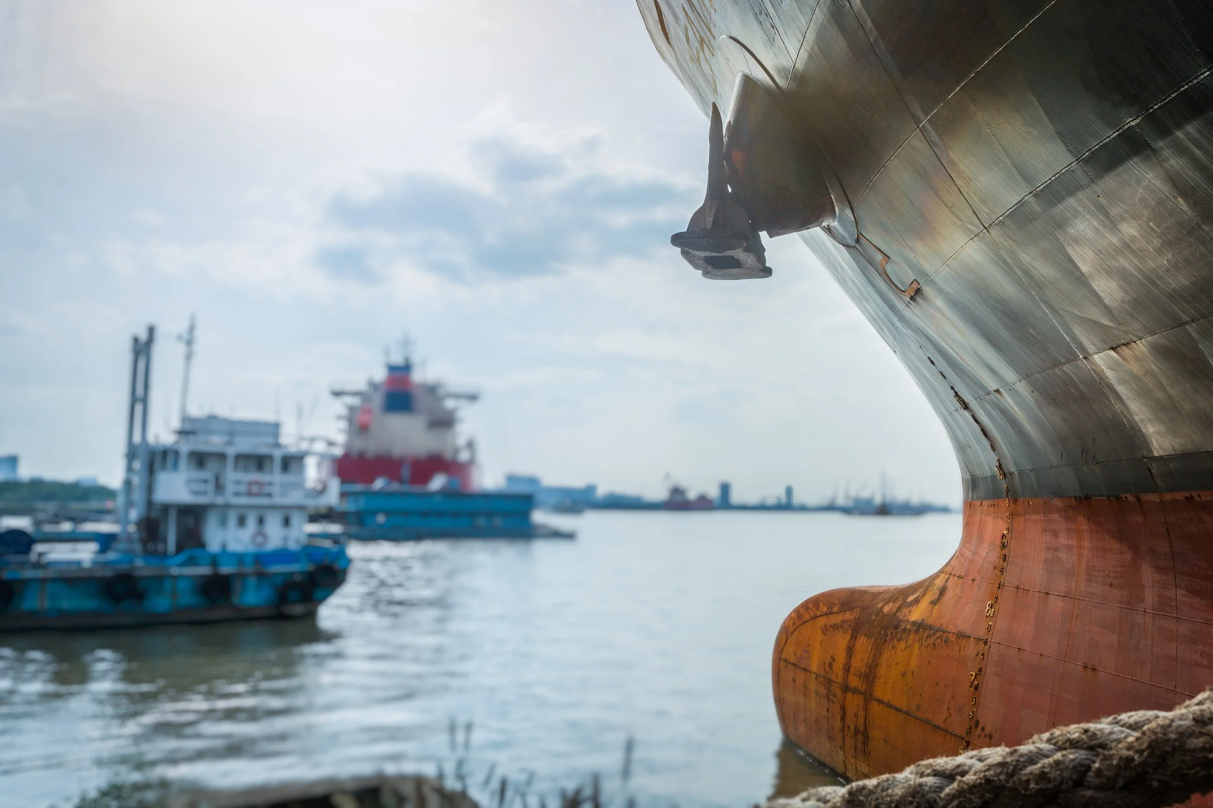 Close-up of a large ship's bow with a rusty hull, docked at a harbor with other boats and ships in the background under cloudy sky.