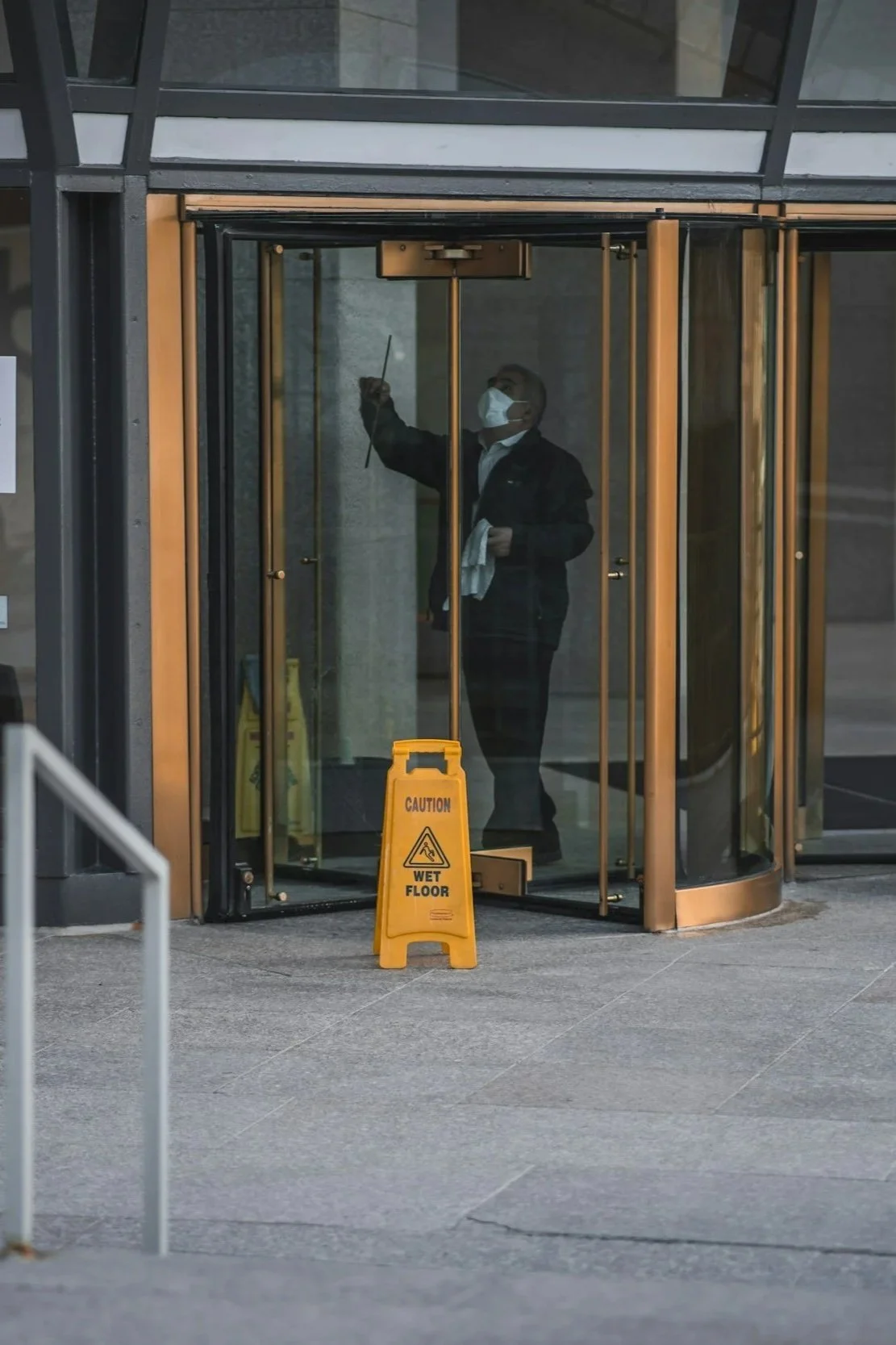 A man wearing a face mask cleaning the glass doors of a revolving entrance, with a yellow caution sign that reads "Caution Wet Floor" in front of him.