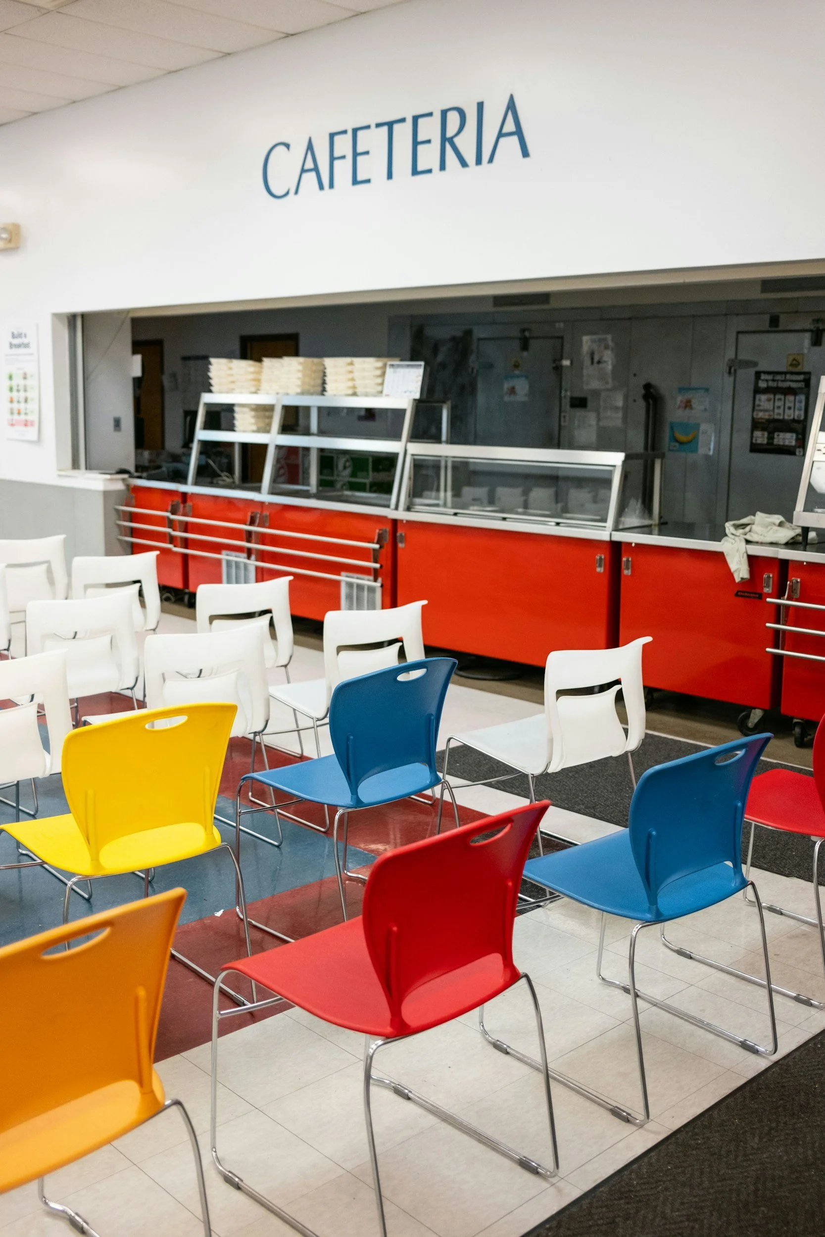 Empty school cafeteria with colorful plastic chairs in yellow, blue, red, and white, arranged in rows on tiled floor with red and white color pattern. Behind the chairs, a serving counter with a sign above that says 'CAFETERIA' in large blue letters is visible.