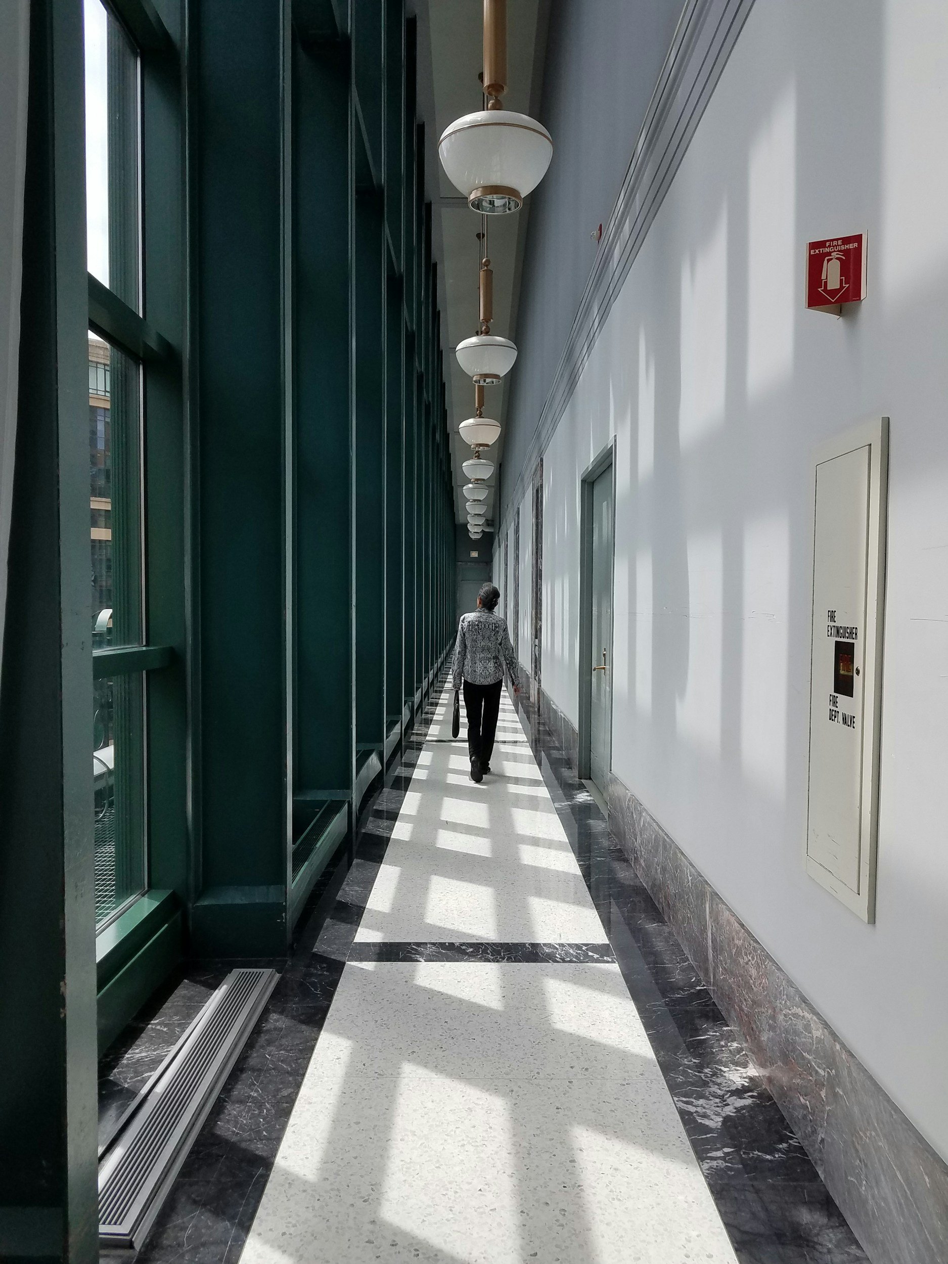 A person walking alone in a bright, narrow corridor with large glass windows on the left and white walls on the right. The corridor has overhead ceiling lights and shadows cast by the windows.