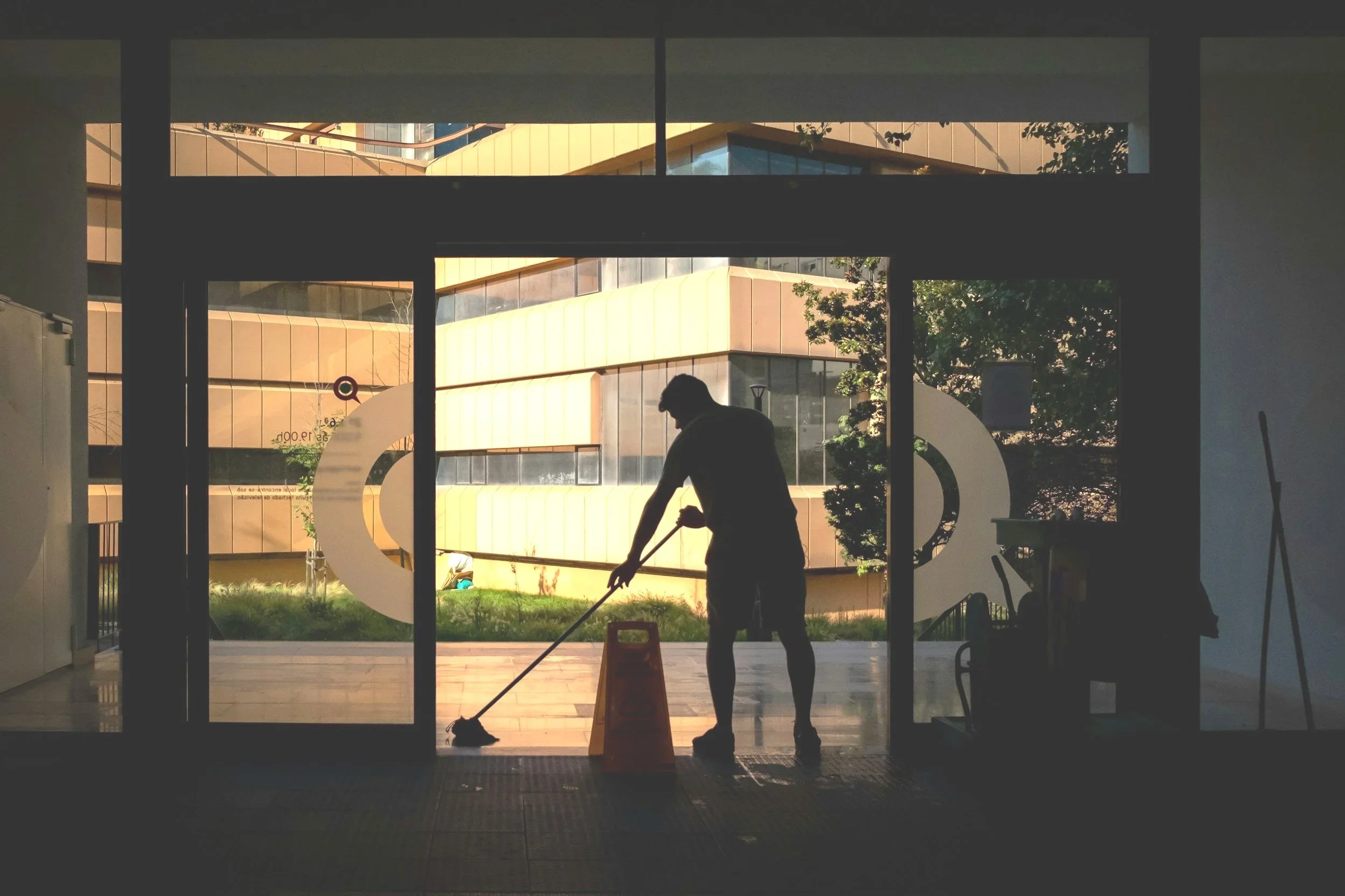 A silhouette of a person mopping the floor inside a building, with large glass doors and windows revealing a modern yellow and brown building and greenery outside.