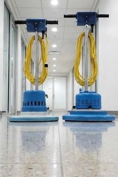 Two floor-cleaning machines with blue bases, yellow hoses, and metal handles in a hallway.