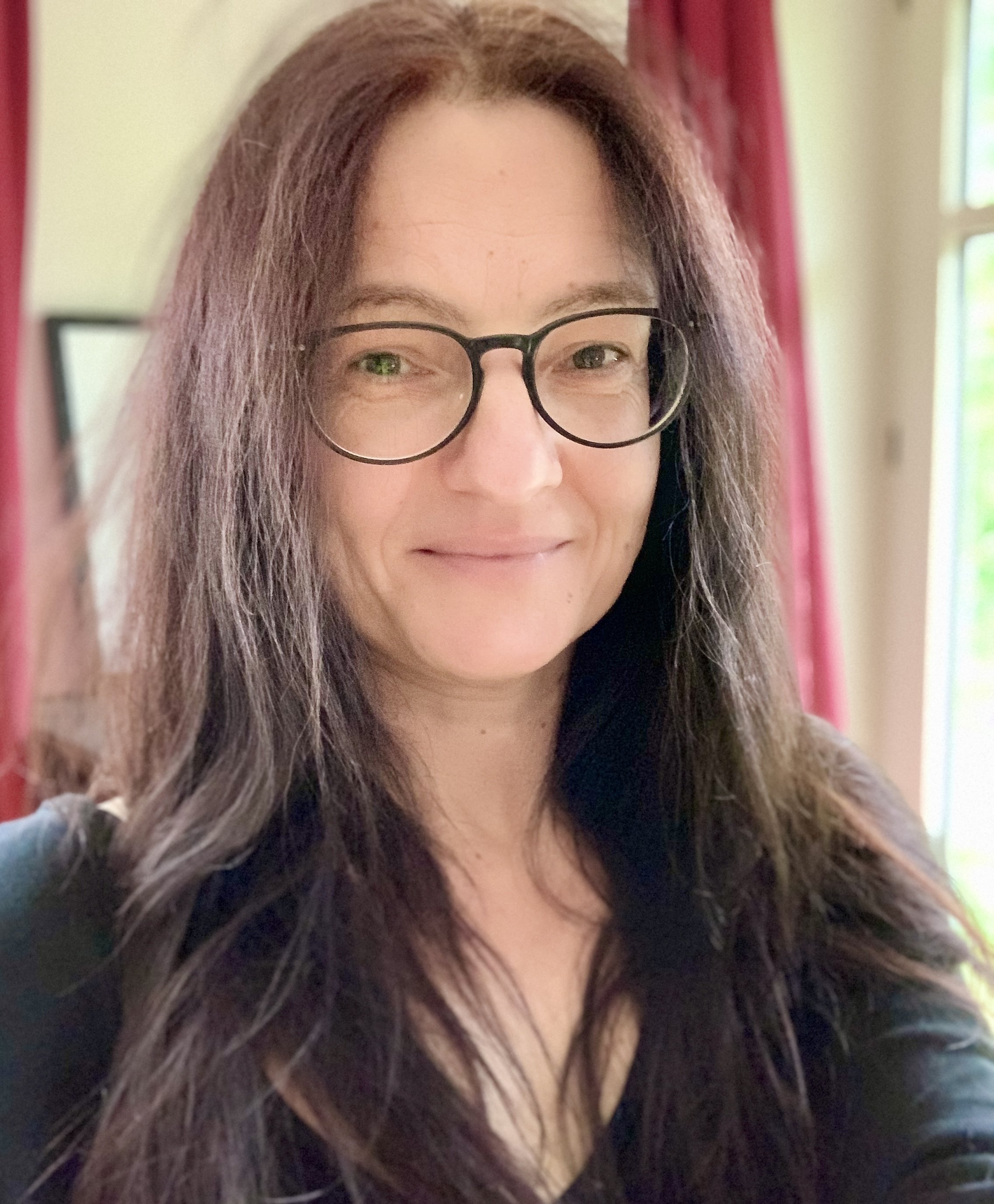 Nahaufnahme der Künstlerin Eva Tenzer mit Brille, lange braune Haare und dunklem Oberteil, im Hintergrund ein Fenster mit roten Vorhängen.