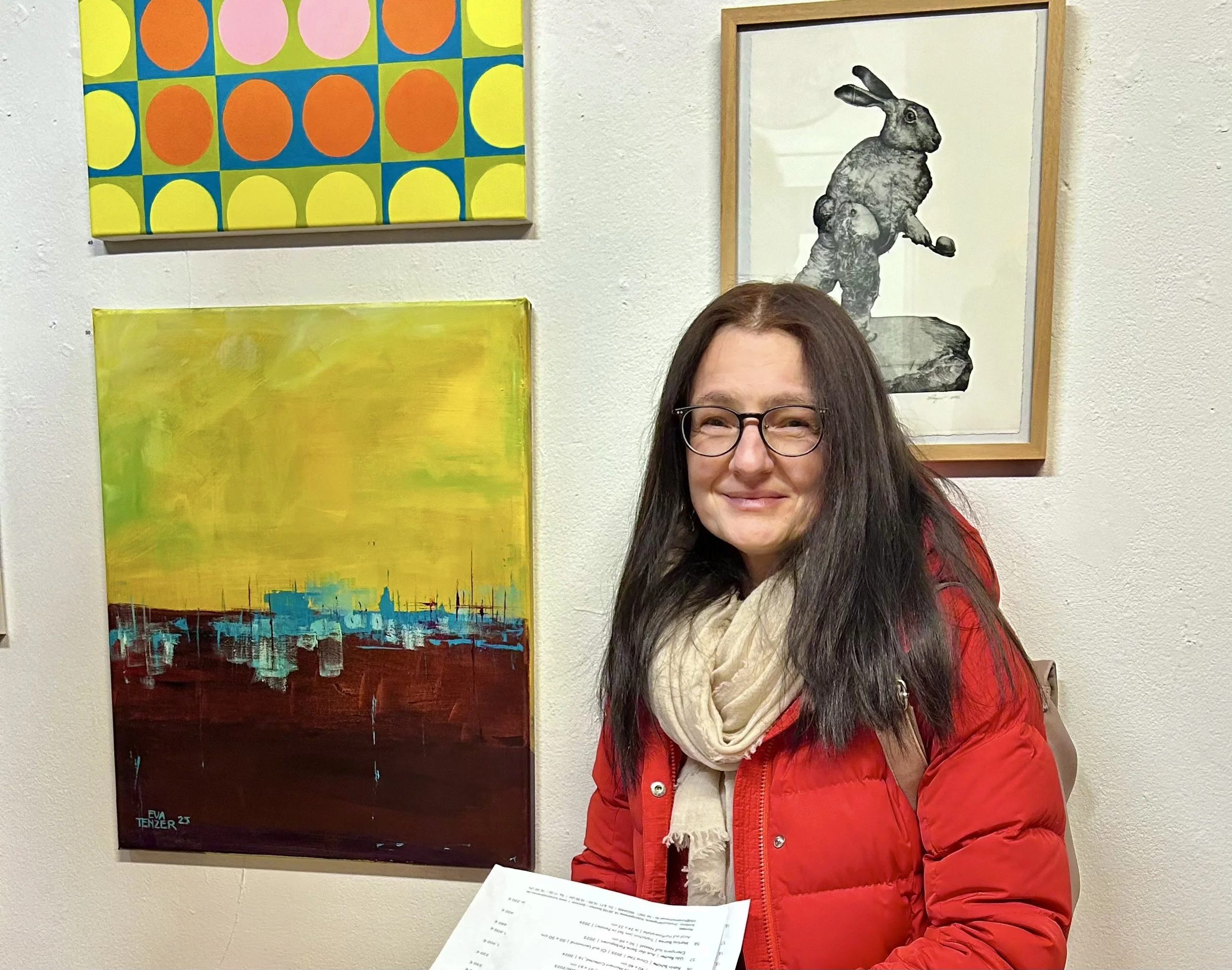 Eine lachende Frau mit schwarzen Haaren, Brille, roter Jacke und einem beige Schal steht vor einer weißen Wand mit bunten Kunstwerken.