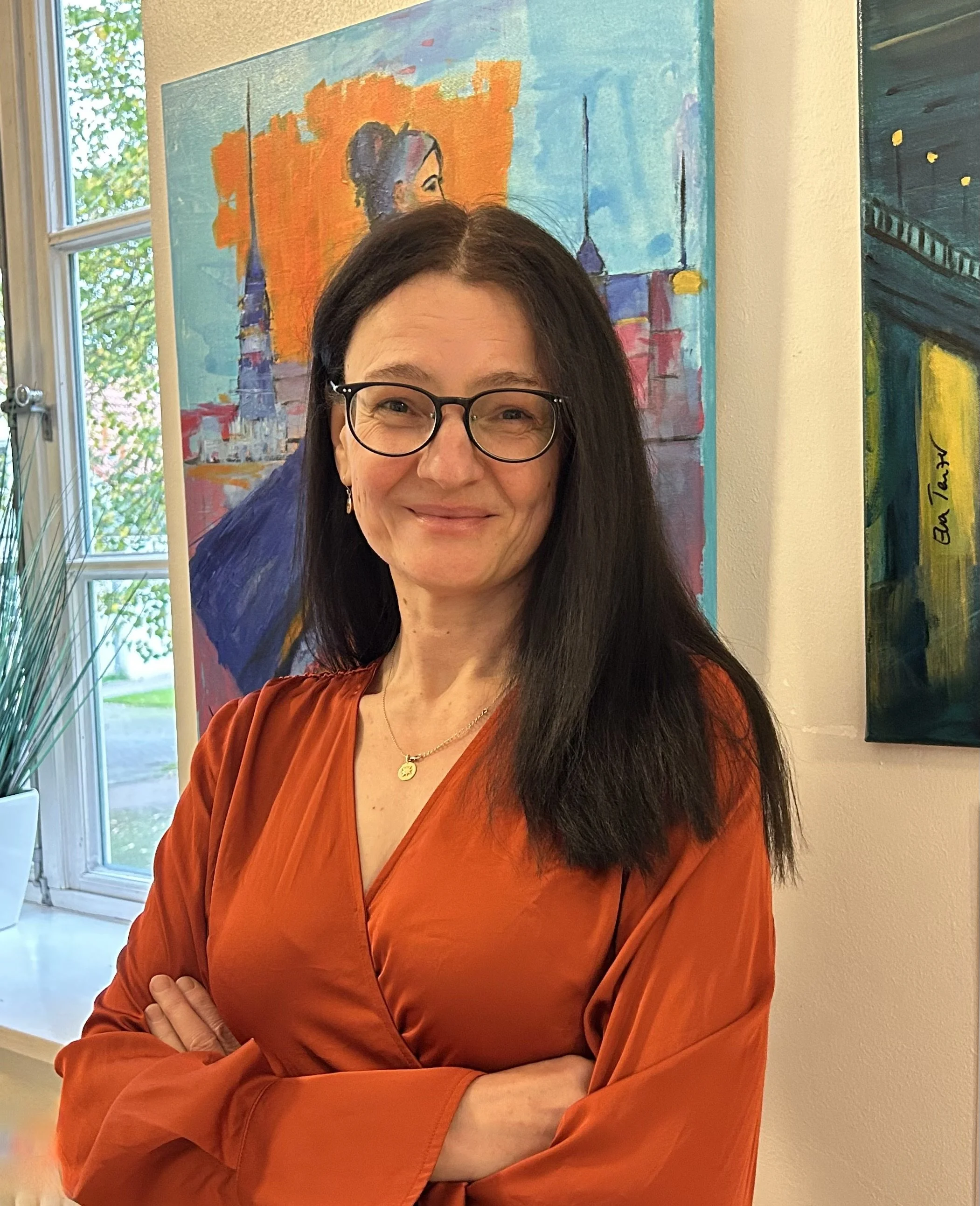 Eine smiling Frau mit schwarzen Haaren, Sonnenbrille und rotem Edelstein, die in einem Kunststudio steht. Sie trägt ein rotes Kleid und steht vor bunten Gemälden, darunter eines mit einer Silhouette einer Frau vor einem orange-blauen Hintergrund.